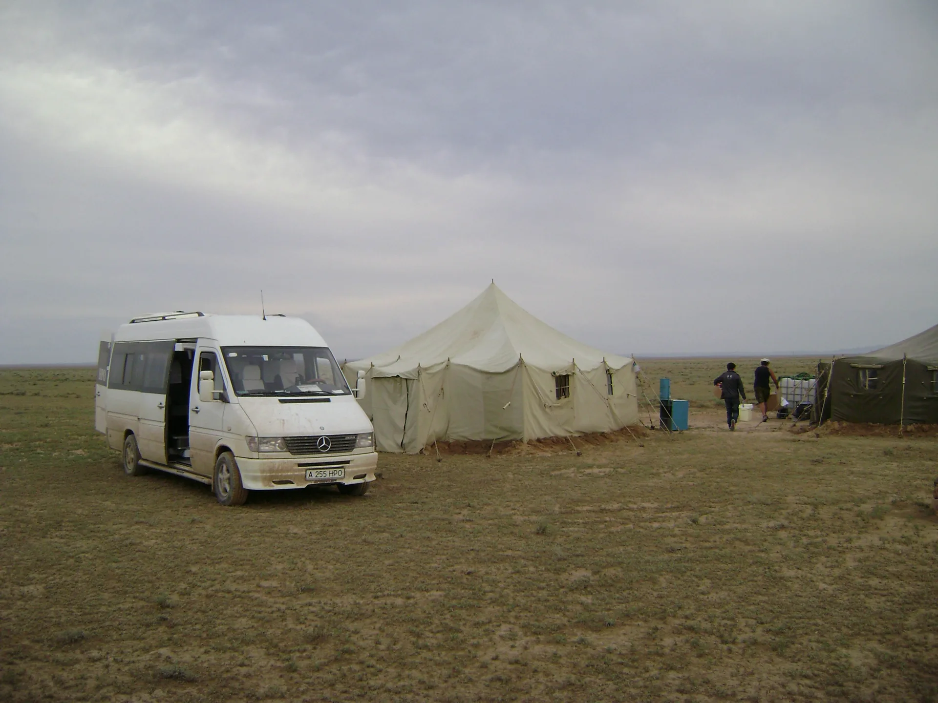 A cluster of traditional Kazakh yurts set on open Taukum Desert sand near the Altyn-Emel National Park boundary, with sandy steppe stretching to the horizon under a blue sky.