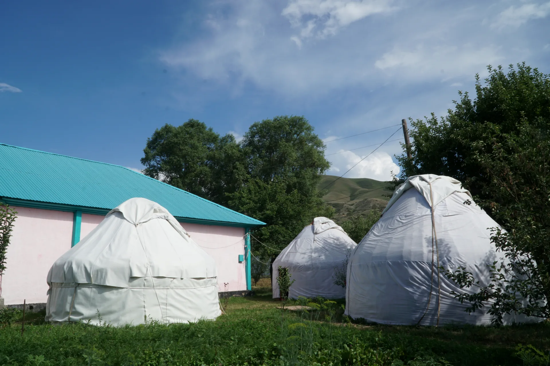 Courtyard of a Kazakh village guesthouse in Saty with two white yurts set up beside a pink-walled building under green trees and a mountain backdrop
