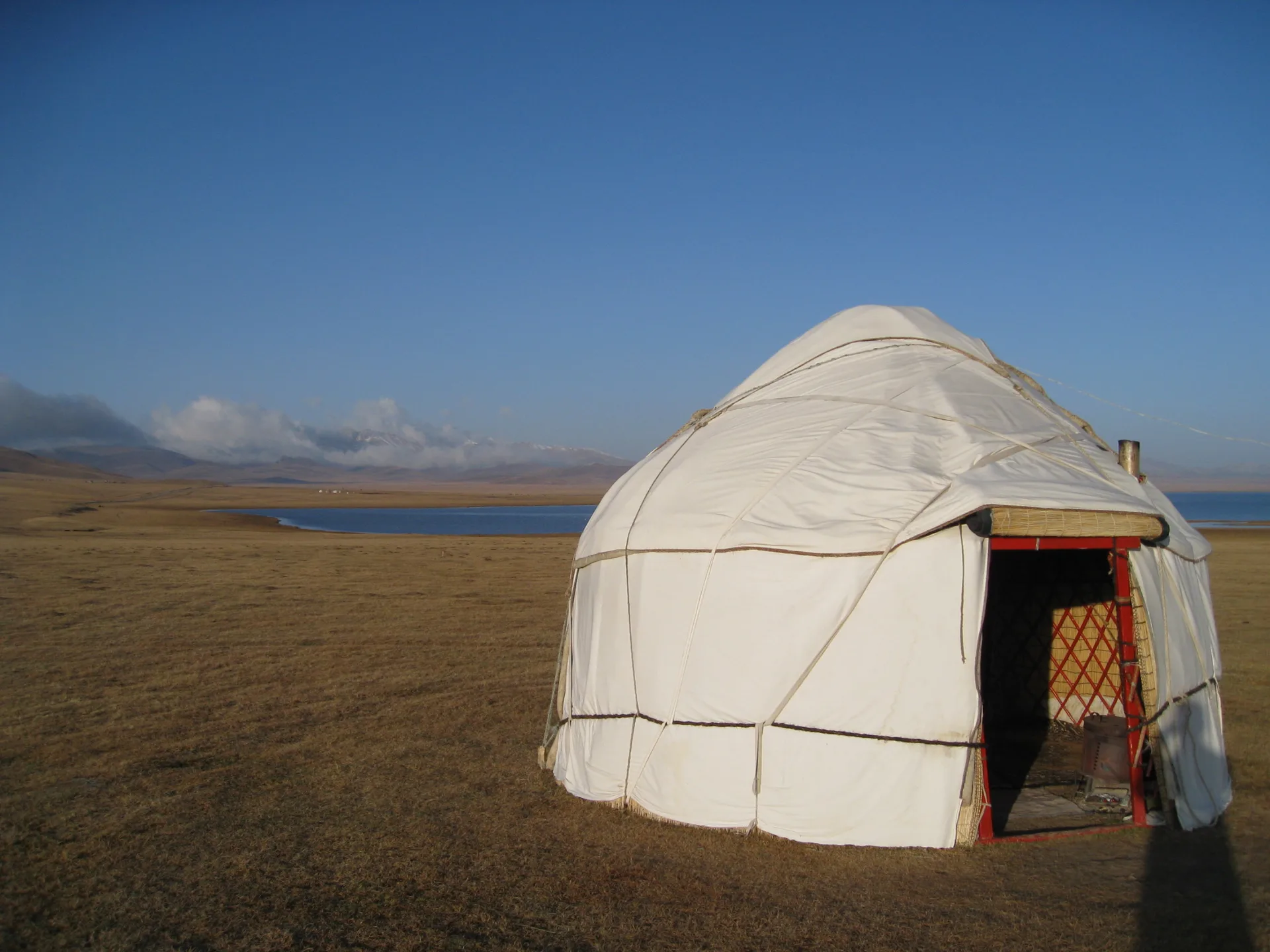 White Kazakh yurt standing alone on open dry steppe at golden hour, with a lake and mountain ridgeline visible in the distance under a clear blue sky