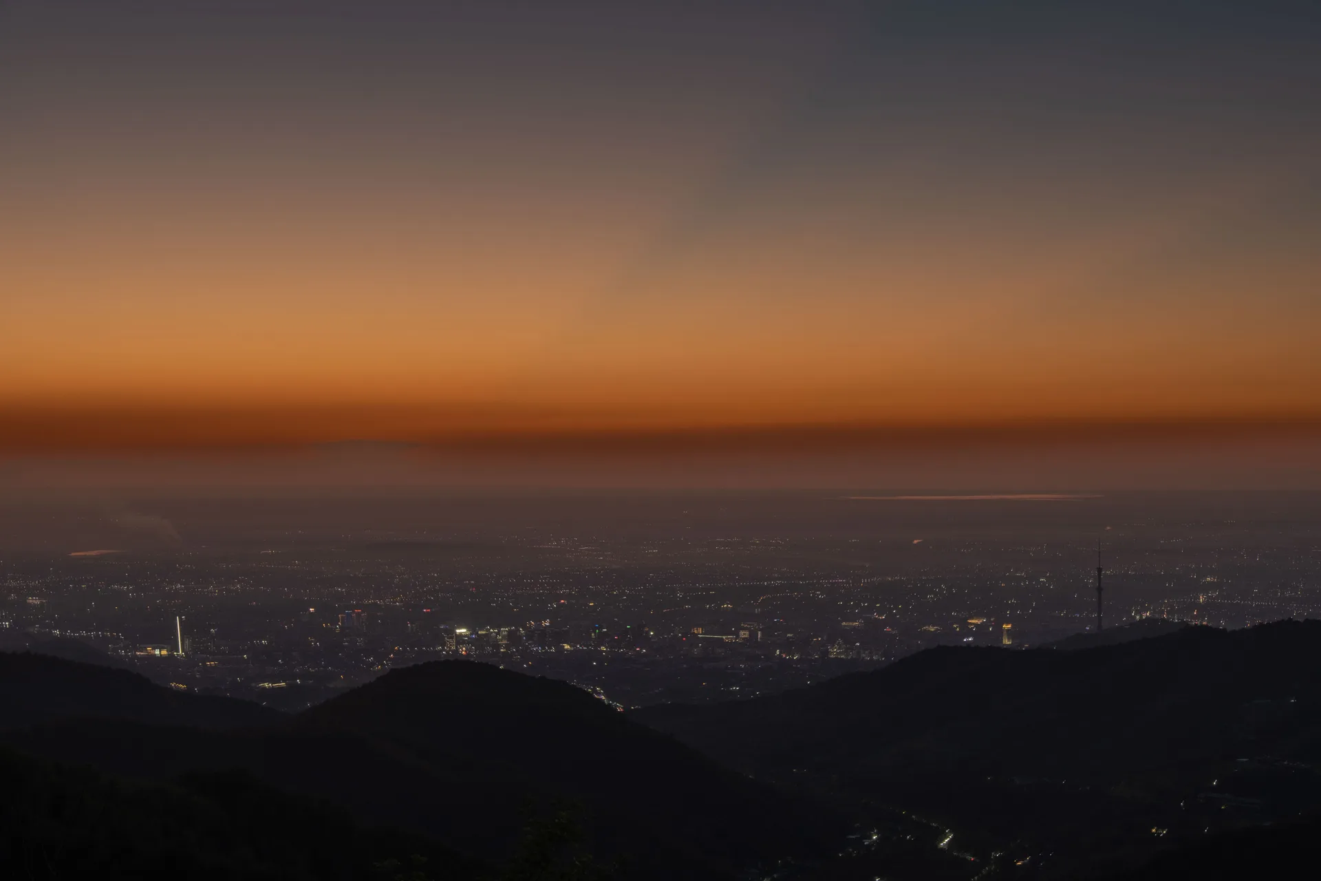Almaty city at evening from a hilltop vantage, high-rise buildings and city lights with the snow-capped Zailiysky Alatau mountains rising sharply behind