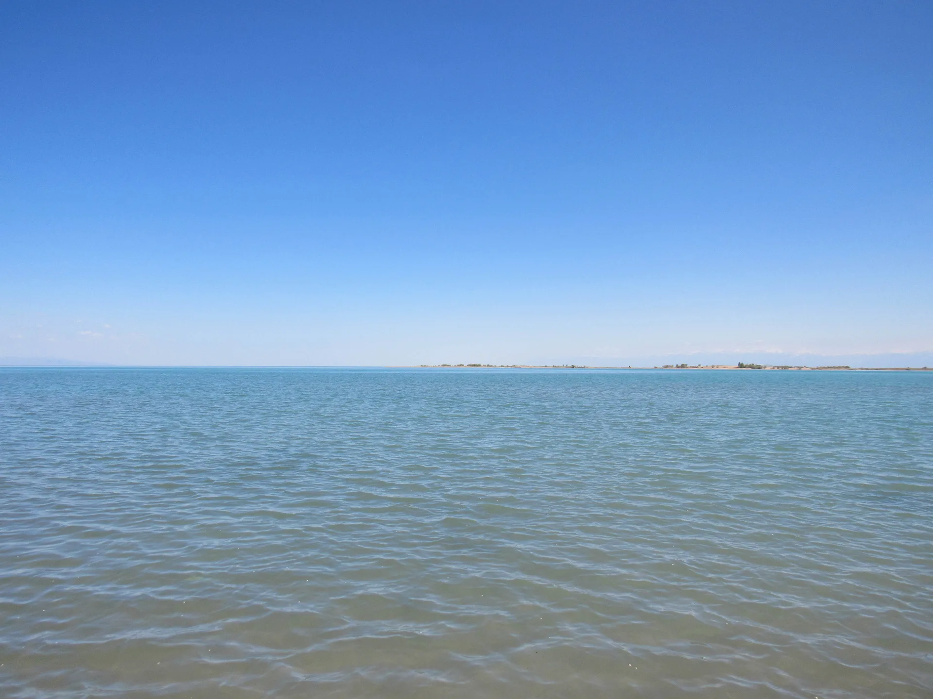 Kapchagay reservoir shoreline at Konaev city, calm blue water with a gravel bank in the foreground under a pale grey sky