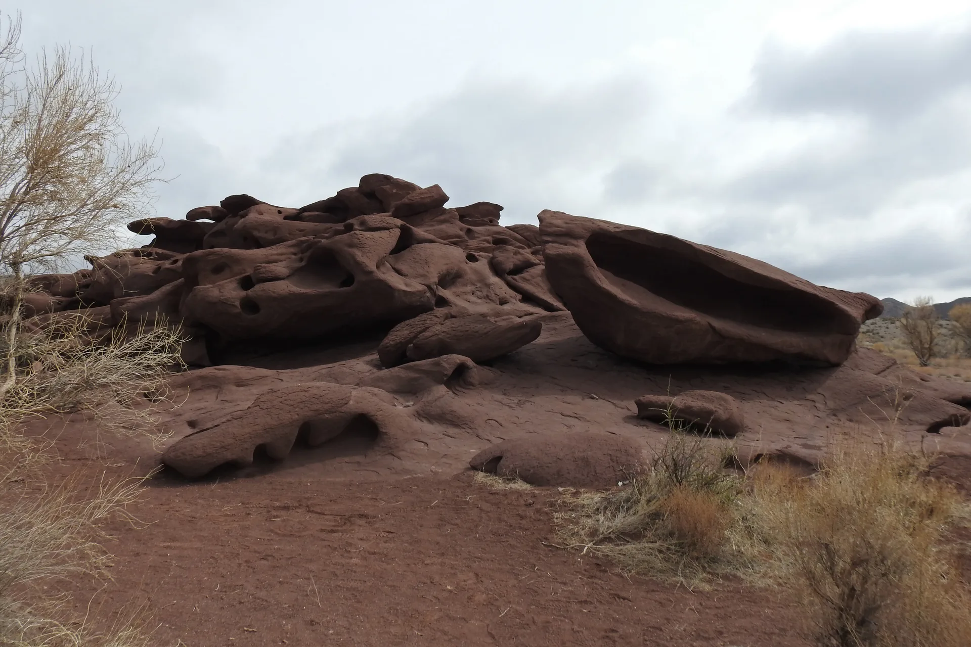 Eroded dark reddish-brown volcanic rock formations of the Katutau Mountains in Altyn-Emel National Park, showing windswept solidified lava outcrops rising above desert scrub under an overcast sky.