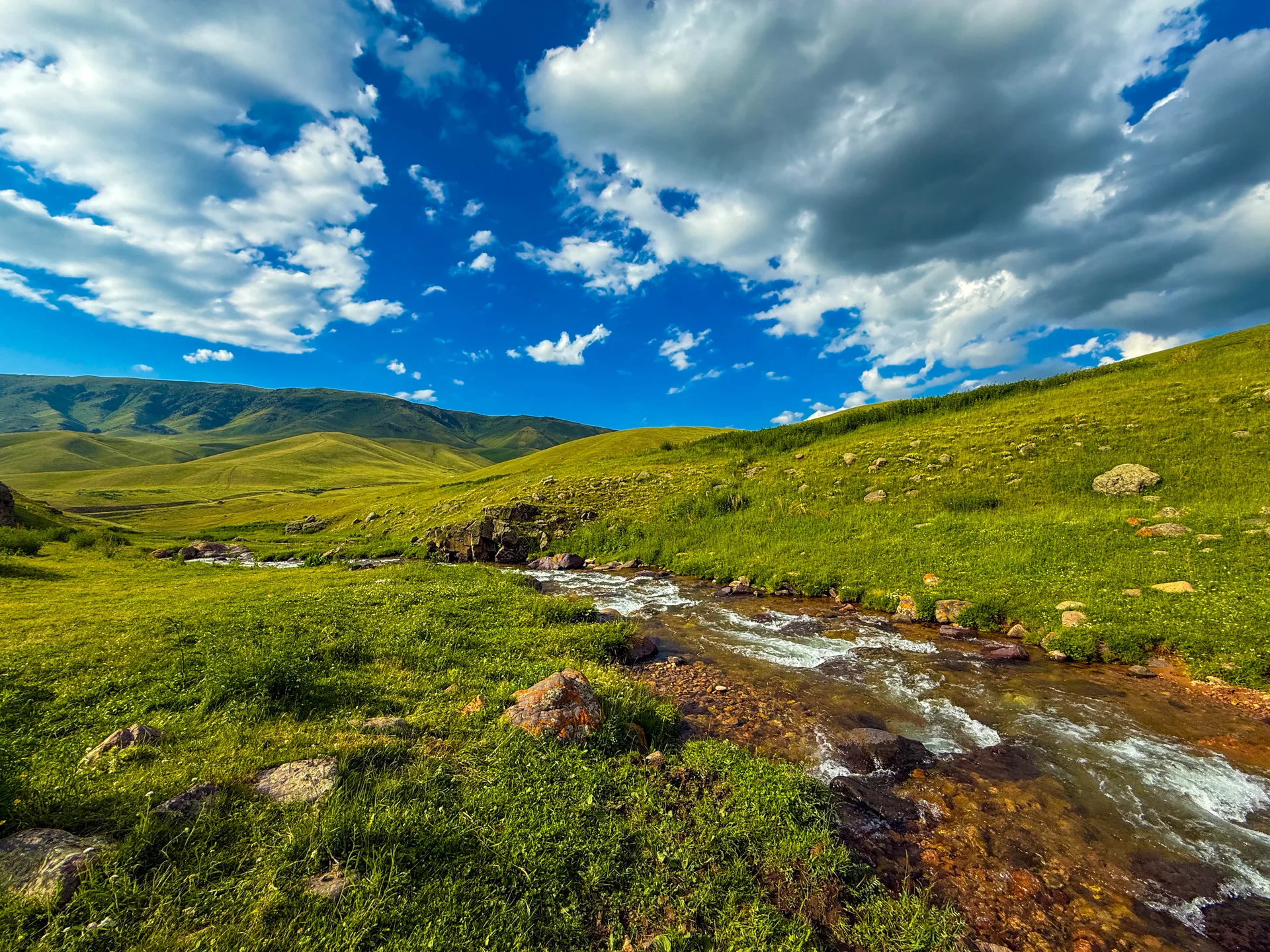 River winding across the floor of Assy Plateau with grassy hillsides rising on both sides under a blue sky.