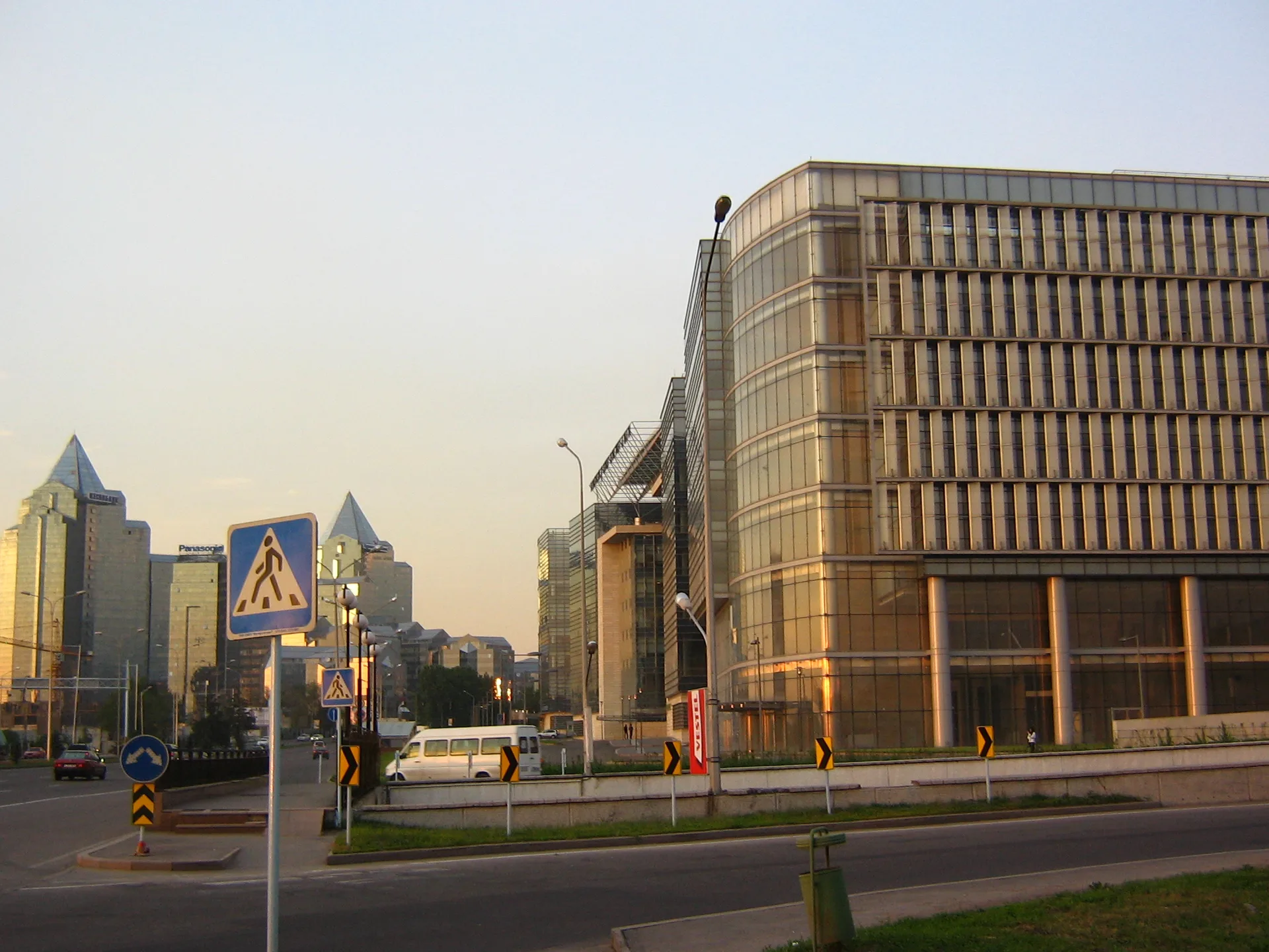 Al-Farabi Avenue in Almaty at dusk, with modern glass skyscrapers lining the road and traffic signs marking the urban departure point.