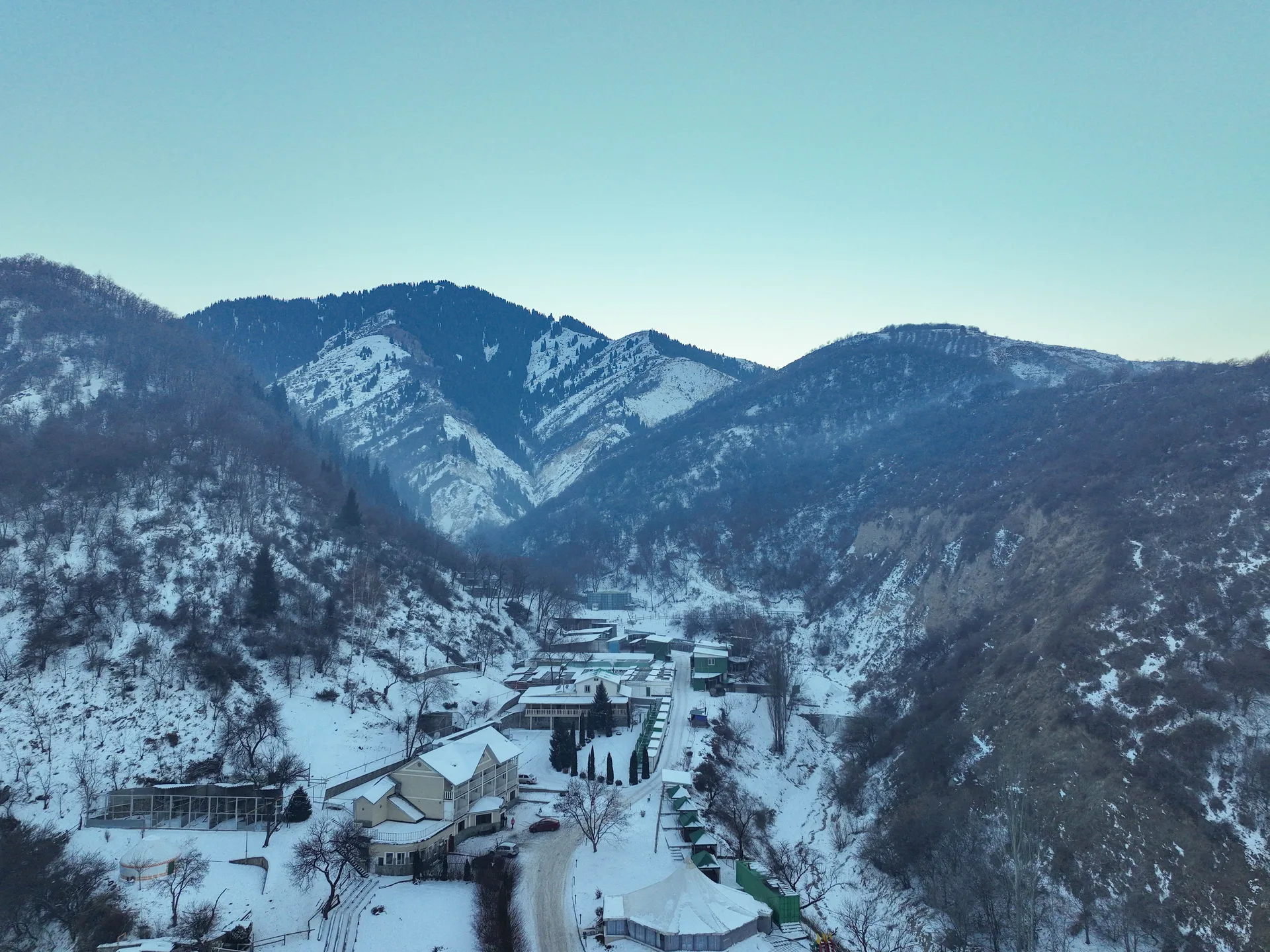 Aerial winter view of the entrance to Big Almaty Gorge (Bolshoe Almatinskoe Ushchelye), showing the gorge mouth settlement and road leading into the snow-dusted mountain valley.