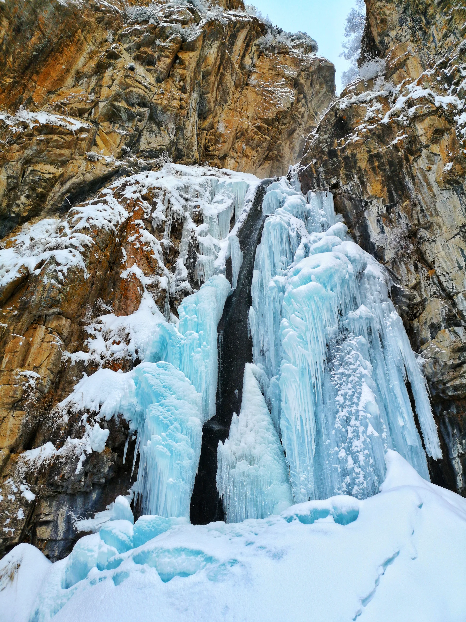 The Lower Butakovka Waterfall frozen into a column of ice in winter, surrounded by snow-covered spruce forest in Ile-Alatau National Park.