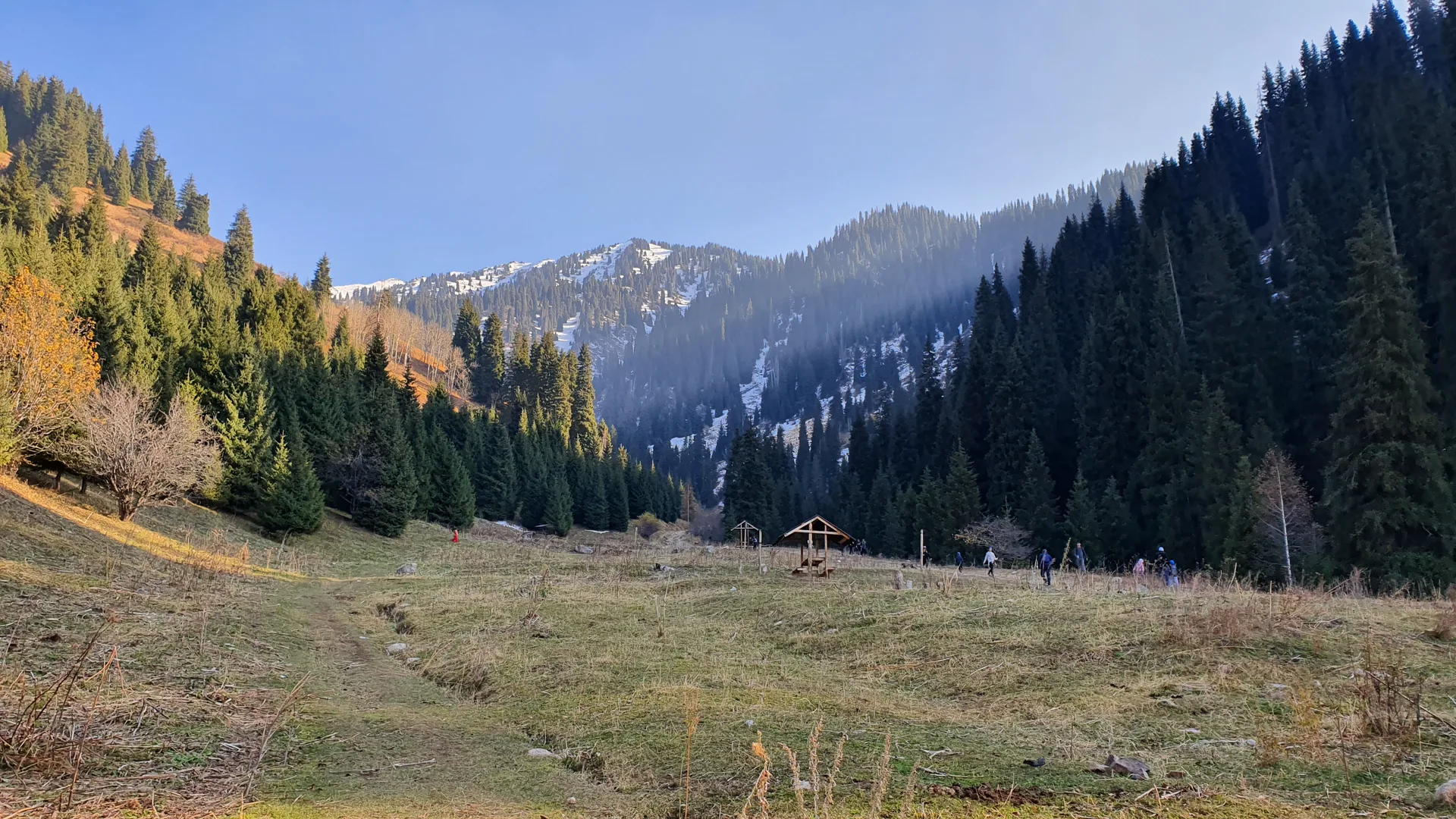 Lower Butakovka Gorge in autumn showing the trailhead area with a wooden shelter, hikers on the path, and dense Tien Shan spruce forest rising to snow-capped peaks in Ile-Alatau National Park.