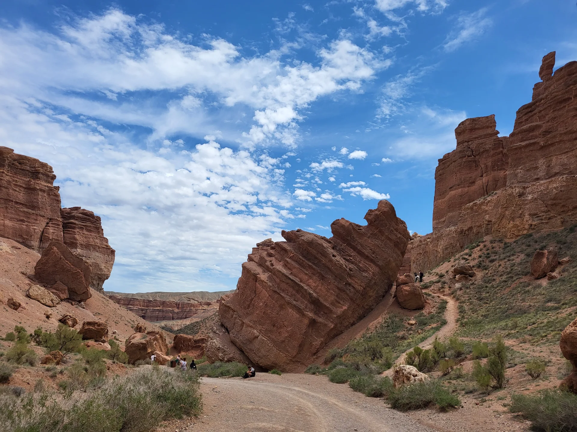 Eroded sandstone formations of Charyn Canyon rising above the canyon floor in shades of red and orange, with a trail visible below.