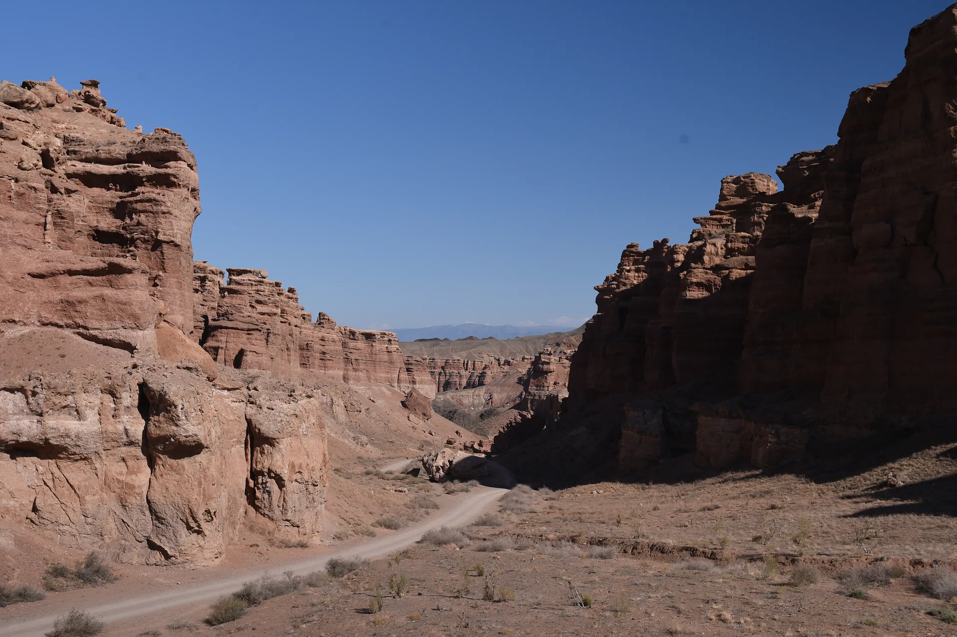A sandy canyon-floor path curves between the 80-metre sandstone towers of Charyn's Valley of Castles, the Tian-Shan foothills visible at the far end of the corridor.