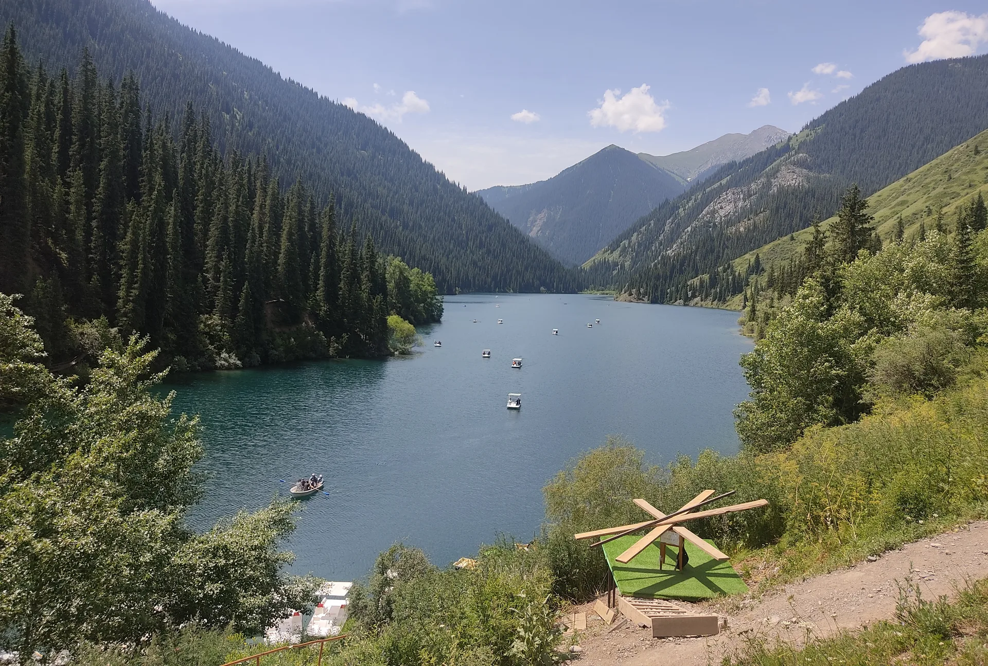 Lower Kolsai Lake at 1,818 m, dense Tian-Shan spruce crowding down to the shoreline and mirrored in calm, deep-green water.