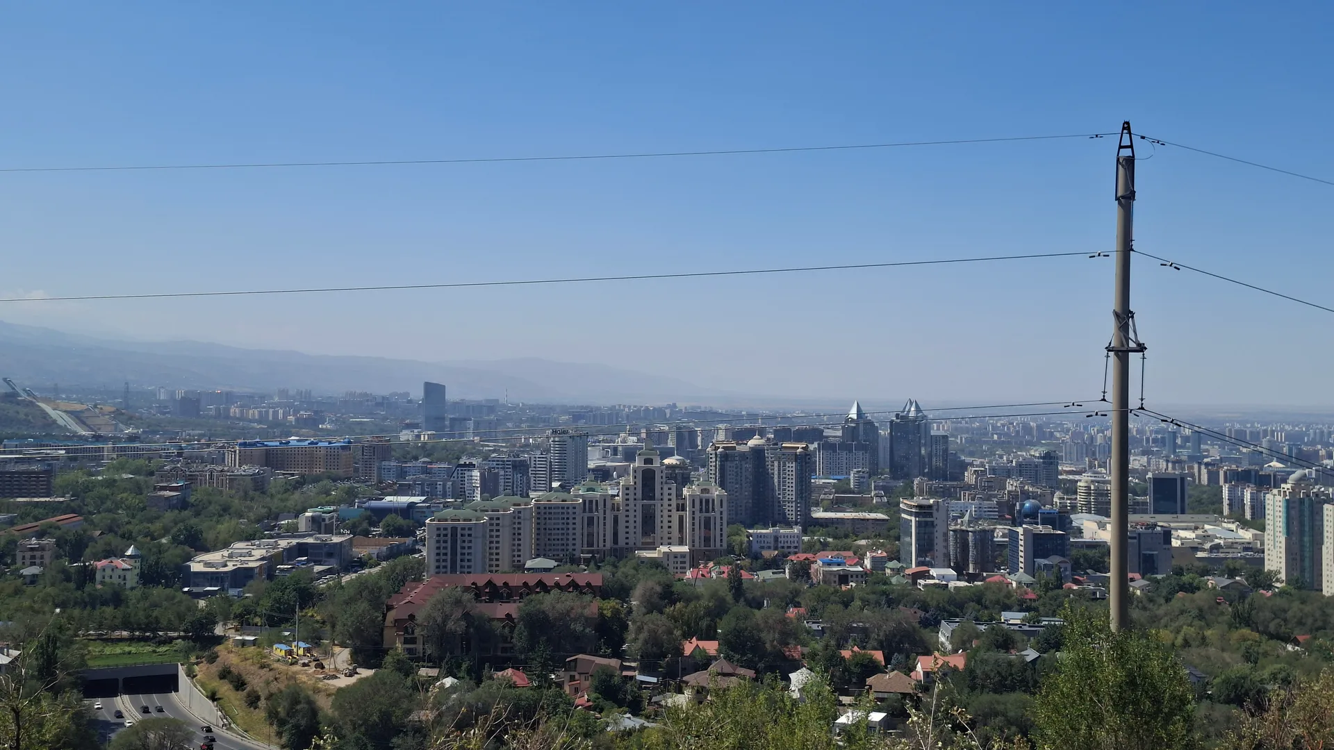 Almaty spreads across a wide valley below Kok Tobe hill, apartment towers and parks backed by the snow-dusted ridgeline of the Zailiysky Alatau under a clear summer morning sky.