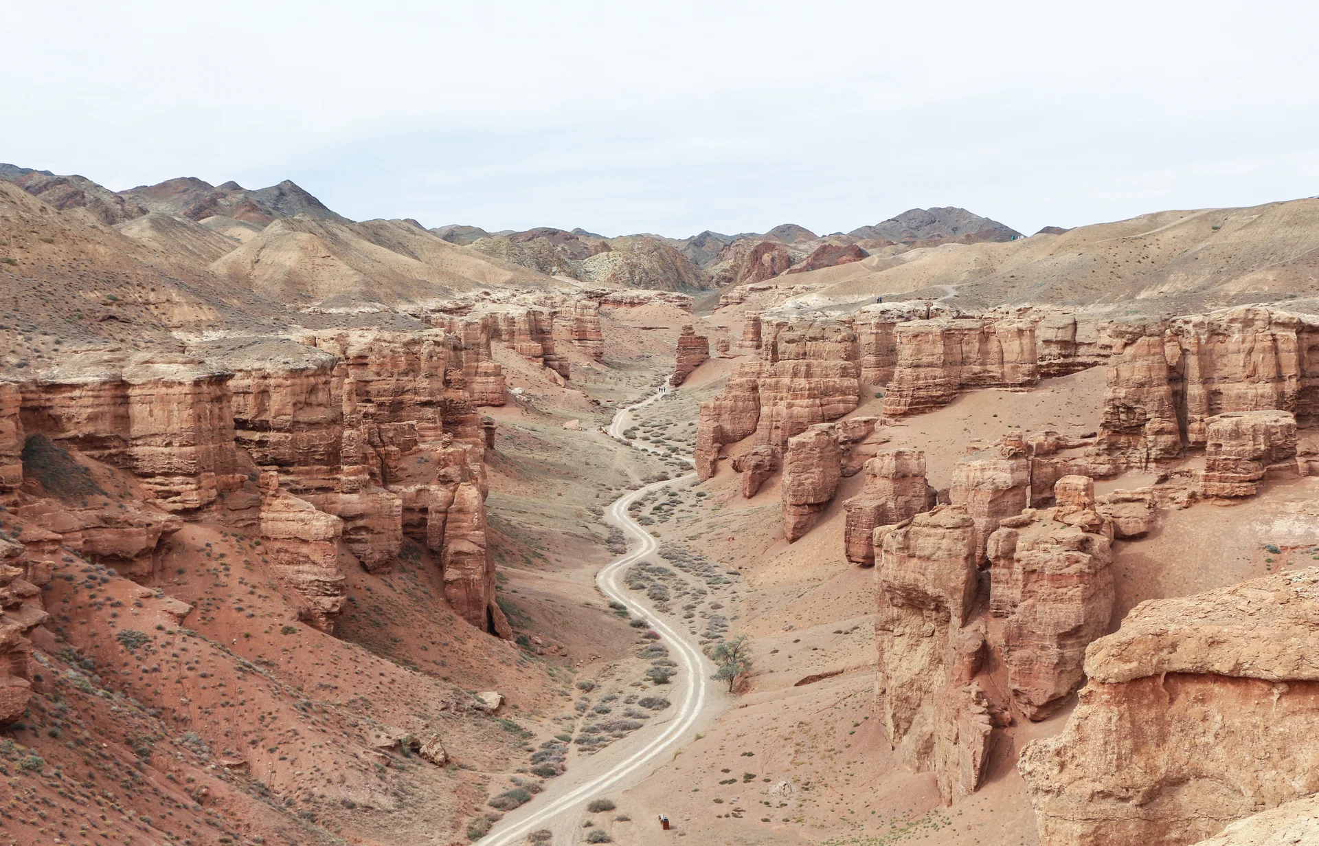 View from the canyon rim looking down into the Valley of Castles: a dirt access road winds between rust-red sandstone towers rising roughly 100 m on both sides, the steppe receding to hazy mountains beyond.