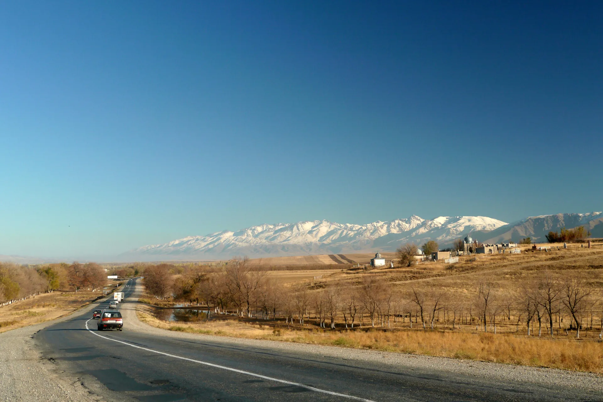 An empty two-lane asphalt road cuts through golden autumn steppe in Kazakhstan, the Tian-Shan range rising faintly on the horizon under a wide blue sky.