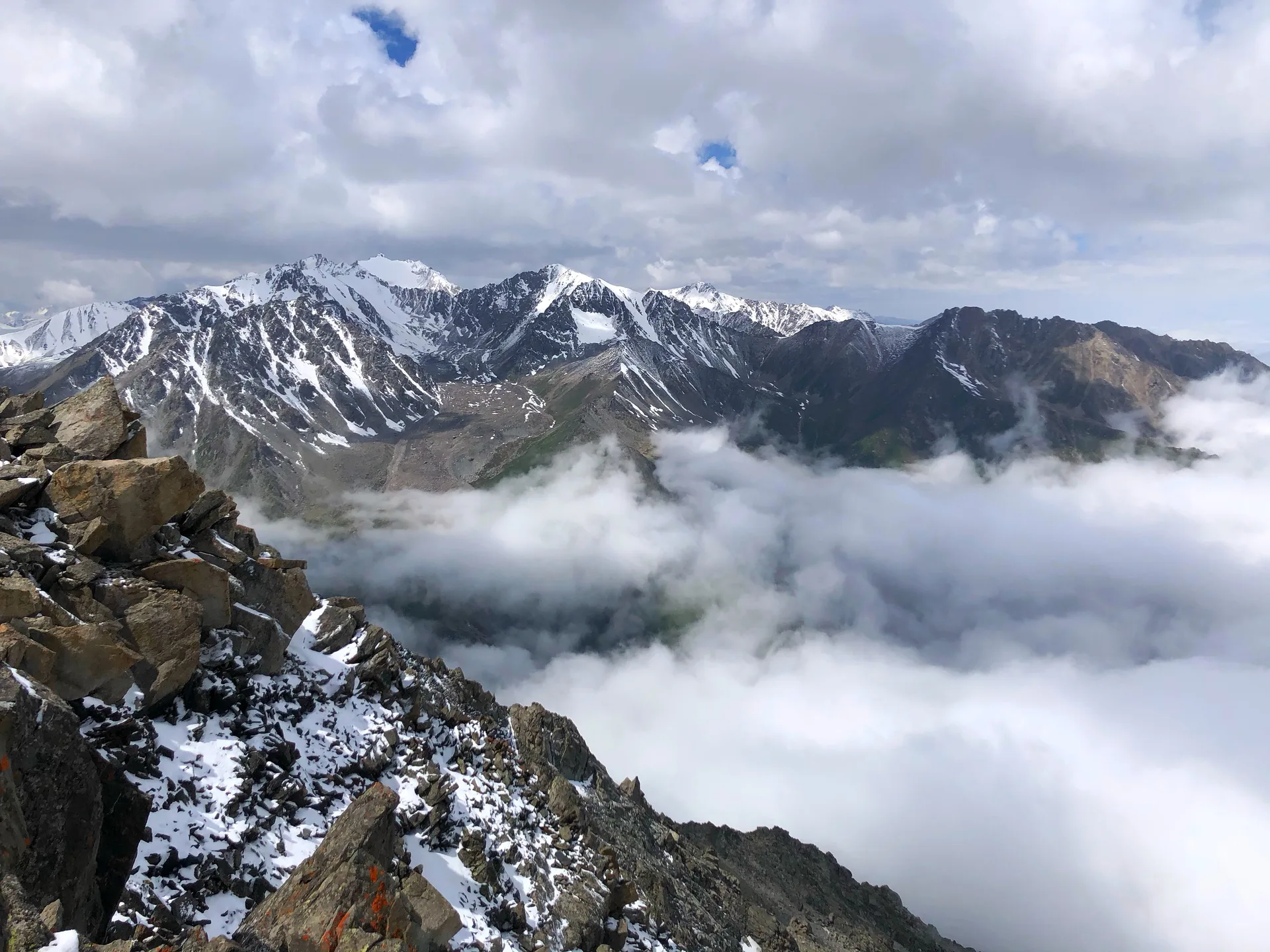 Rocky summit ridge in Ile-Alatau National Park with snow-patched Trans-Ili Alatau peaks and clouds filling the valleys below, viewed from a high-alpine summit above 3,000 m.