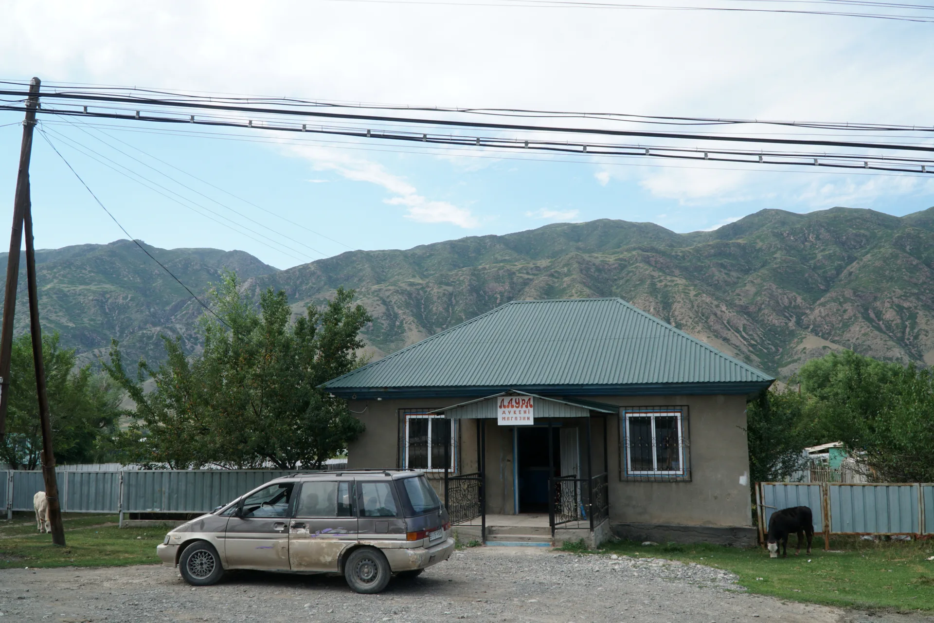 A guesthouse in Saty village with a car parked outside and the green Tien Shan foothills rising behind, Almaty Region, Kazakhstan.