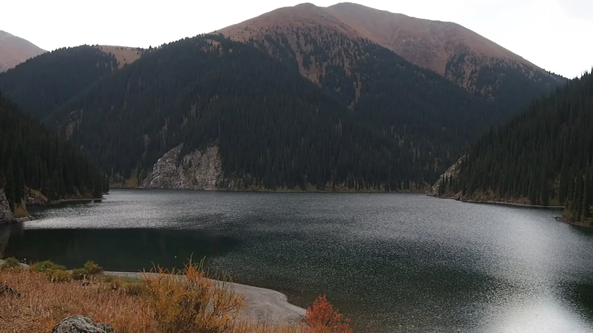 The second (middle) Kolsai Lake in autumn, a wide alpine lake enclosed by spruce-covered slopes and a high rocky ridge under overcast skies.
