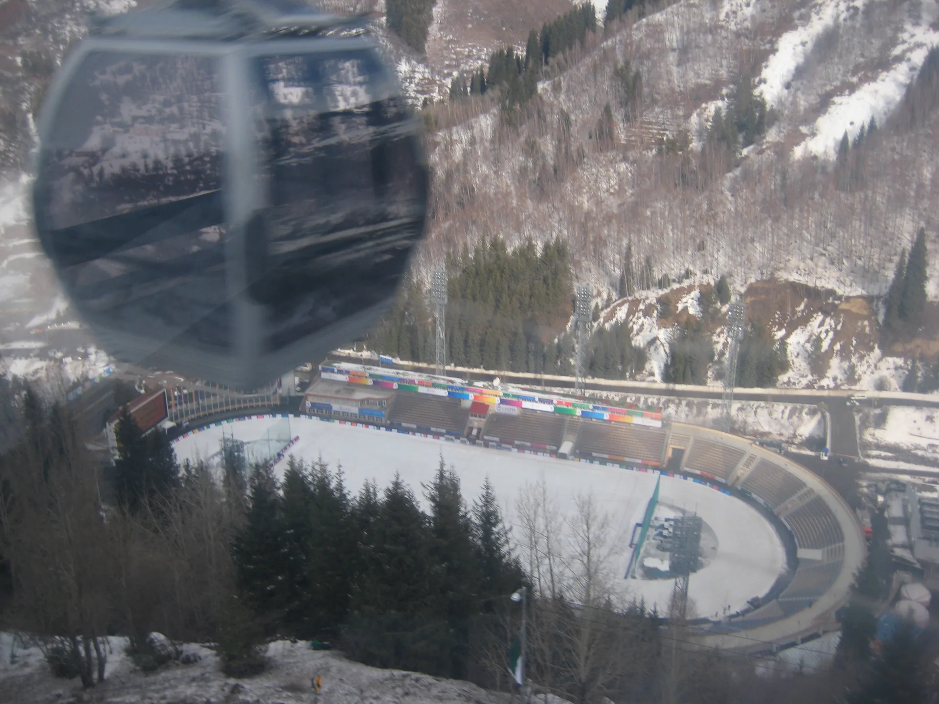 Aerial view of Medeu ice rink stadium and the surrounding gorge, photographed from the height of the Shymbulak gondola cable car, showing the stadium's expanse of ice and the protective dam ridge behind it.