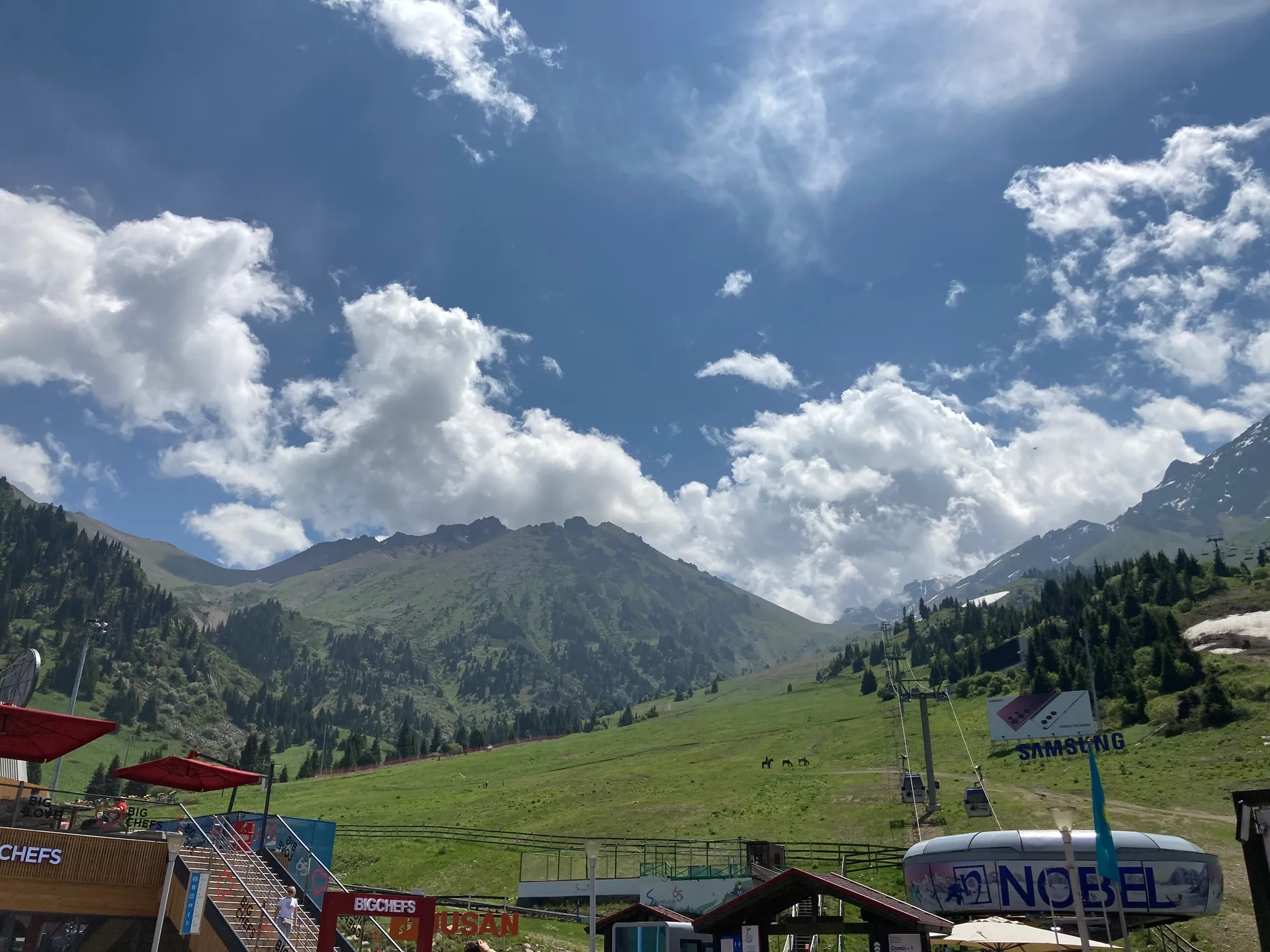 Shymbulak ski resort base station at 2,260 m in summer, with gondola terminal buildings, chairlift infrastructure, restaurant terraces, and green alpine meadows backed by the Zailiisky Alatau peaks.