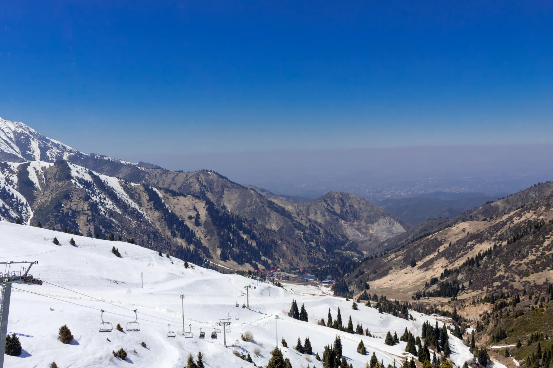 View from Talgar Pass at 3,200 m looking down the snow-covered Shymbulak ski slopes toward the Almaty gorge below, with ski lift pylons, bare rocky ridges, and the city haze visible in the far distance.