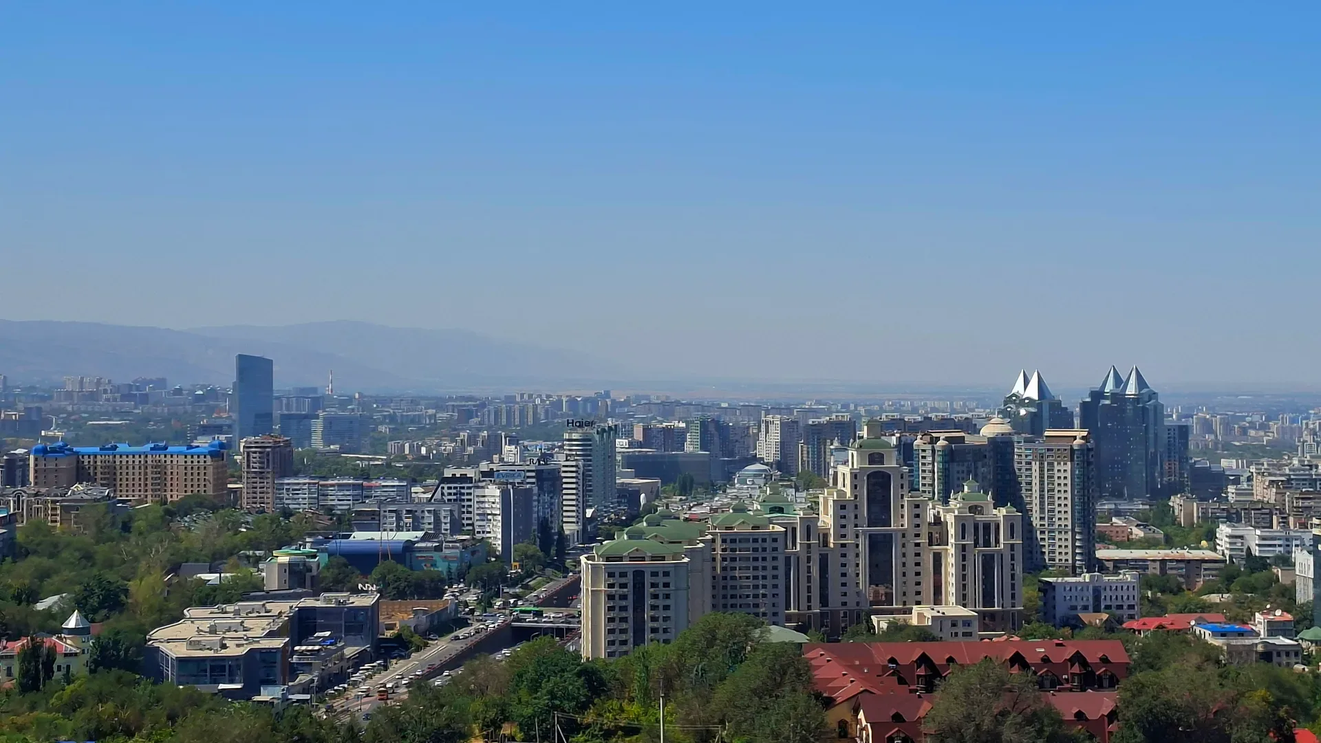 Almaty city skyline viewed from Kok Tobe hill, mid-rise buildings and leafy avenues backed by snow-capped Zailiysky Alatau ridgeline