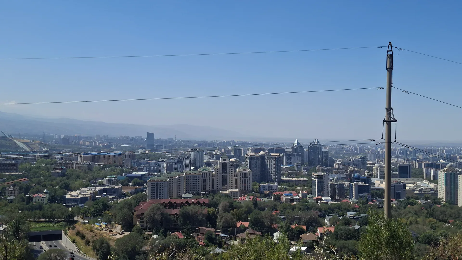 Almaty spreads across a wide valley below Kok Tobe hill, apartment towers and parks backed by the snow-dusted ridgeline of the Zailiysky Alatau under a clear summer morning sky.