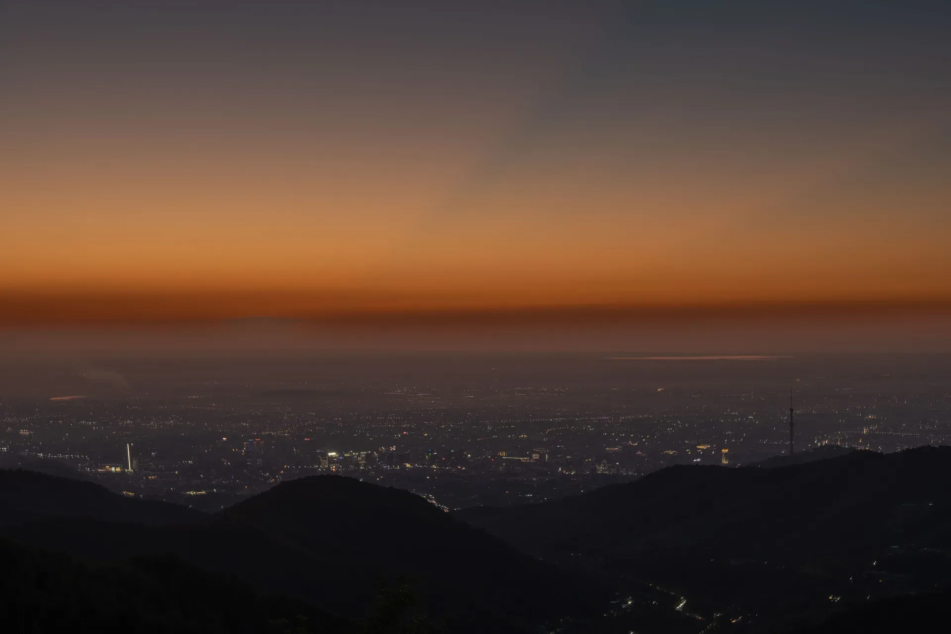Almaty city at evening from a hilltop vantage, high-rise buildings and city lights with the snow-capped Zailiysky Alatau mountains rising sharply behind