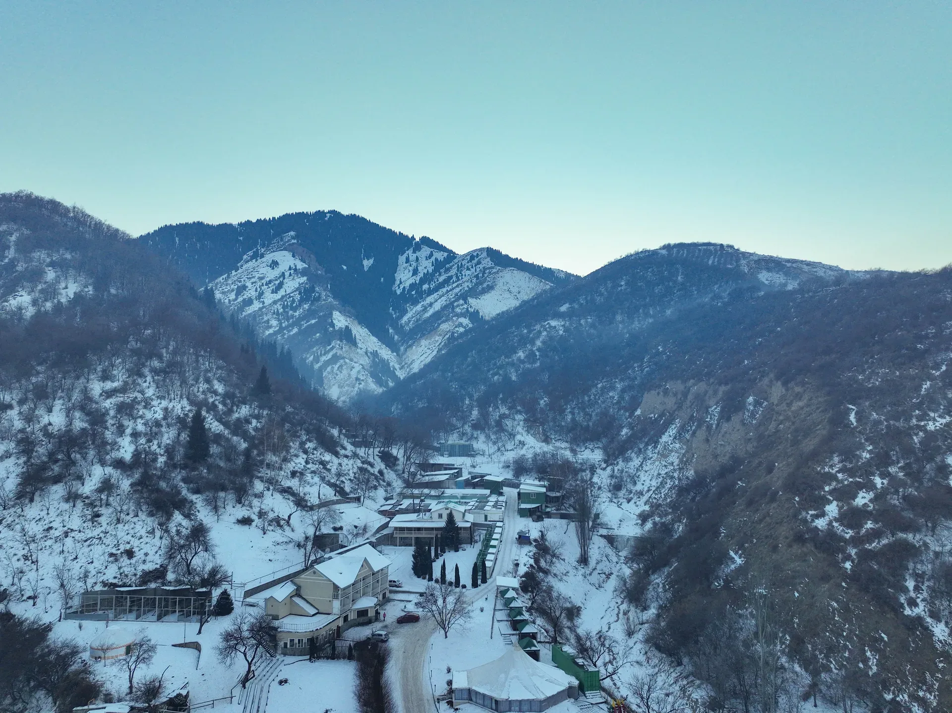 Aerial winter view of the entrance to Big Almaty Gorge (Bolshoe Almatinskoe Ushchelye), showing the gorge mouth settlement and road leading into the snow-dusted mountain valley.