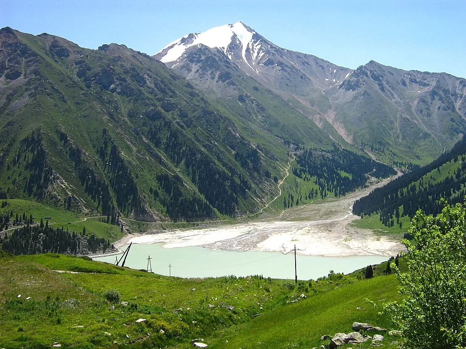June shoreline of Big Almaty Lake with turquoise water below bare mountain slopes.
