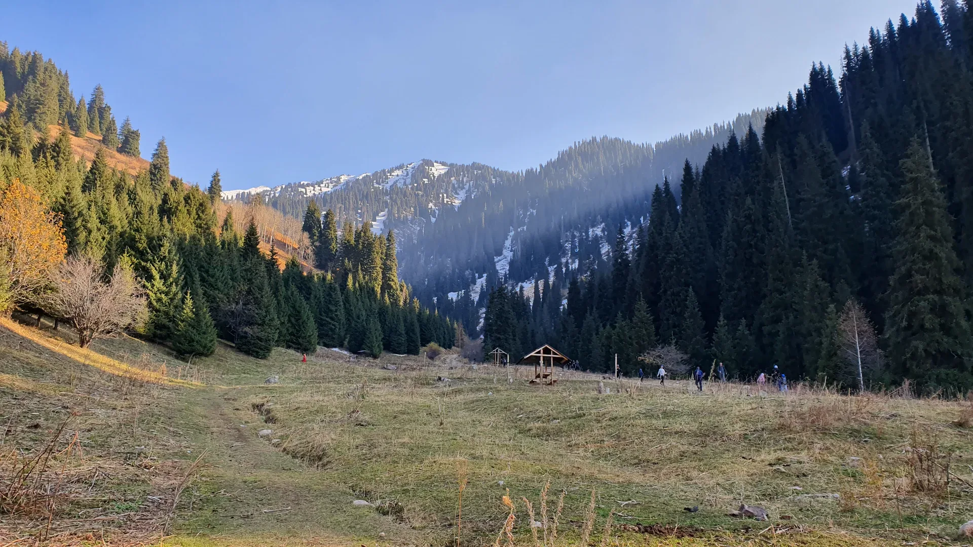 Lower Butakovka Gorge in autumn showing the trailhead area with a wooden shelter, hikers on the path, and dense Tien Shan spruce forest rising to snow-capped peaks in Ile-Alatau National Park.