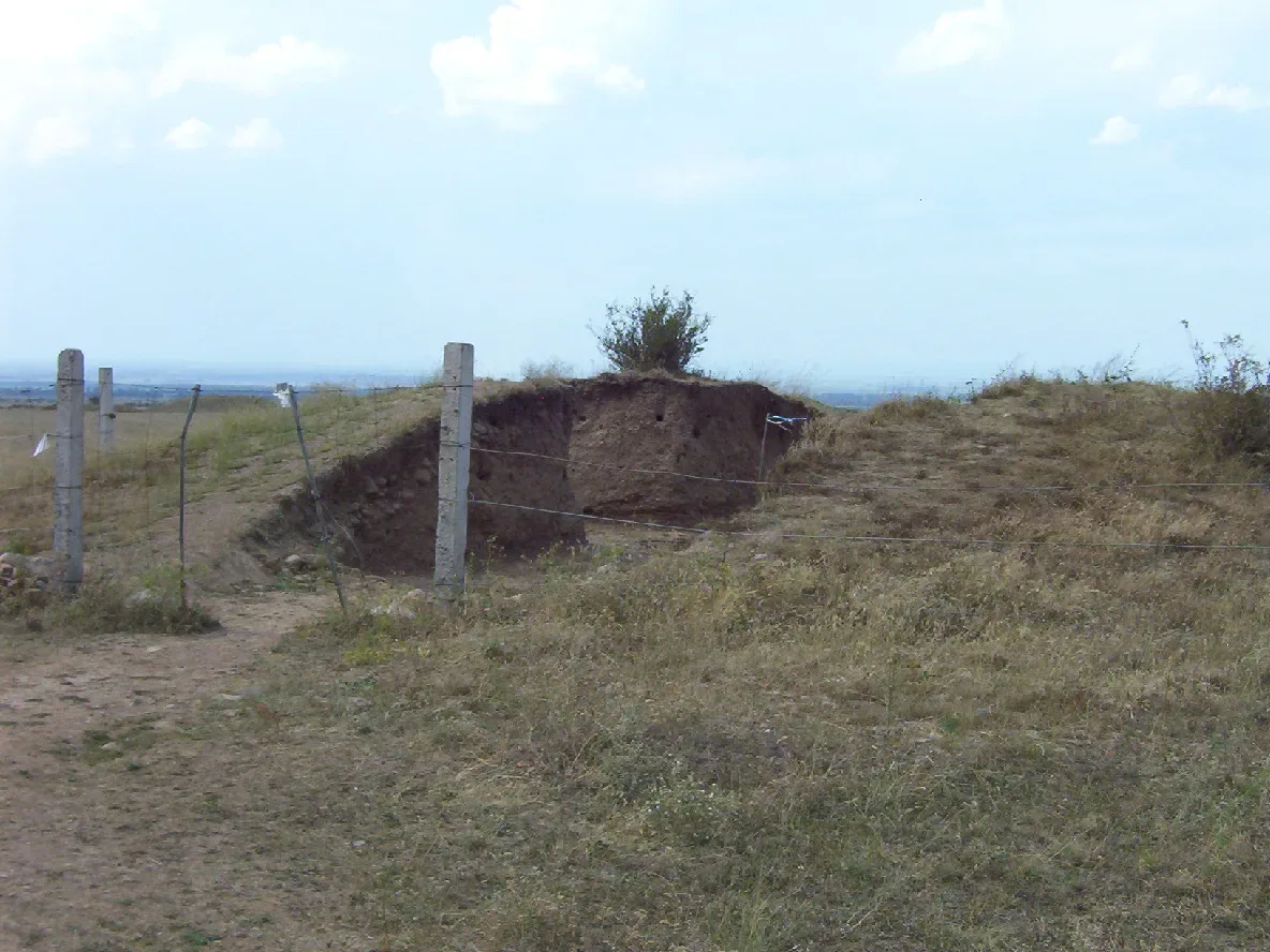 Grassed-over Saka royal burial mound at the Issyk kurgan site in Kazakhstan, fenced with a low rail on open steppe with the Trans-Ili Alatau mountains behind