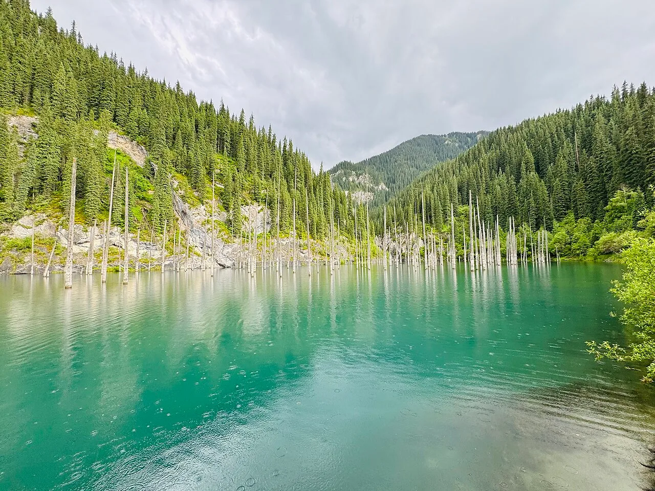 Cloudy Kaindy Lake after rain, showing wet forest slopes near the end of the parking-area footpath.
