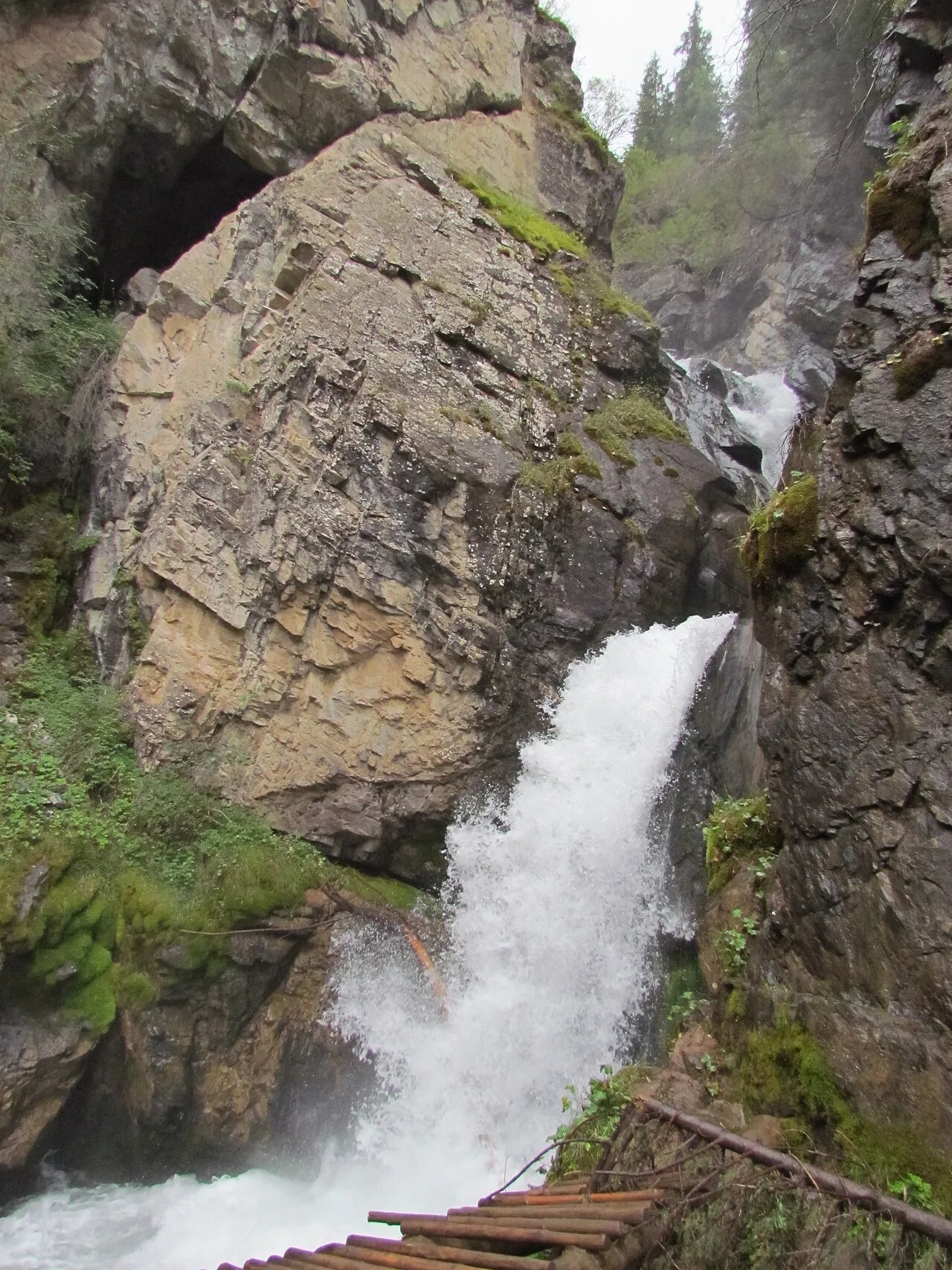 Tall Kairak Waterfall dropping through a narrow rocky cleft with dark wet walls and forest above.