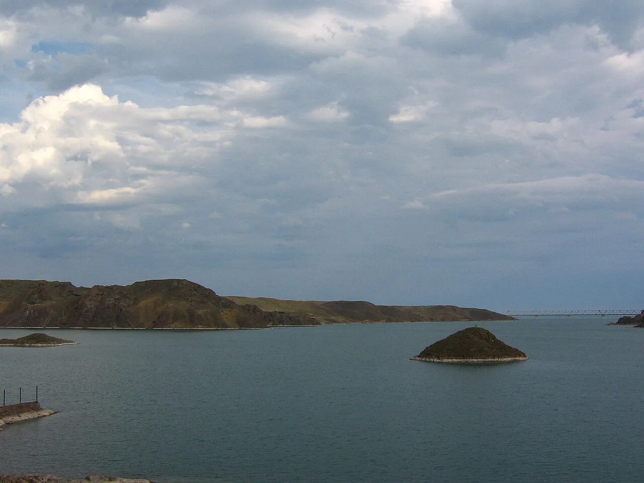 Low sandy bank and roadside view near Kapchagai reservoir, with sparse steppe vegetation beside the crossing.