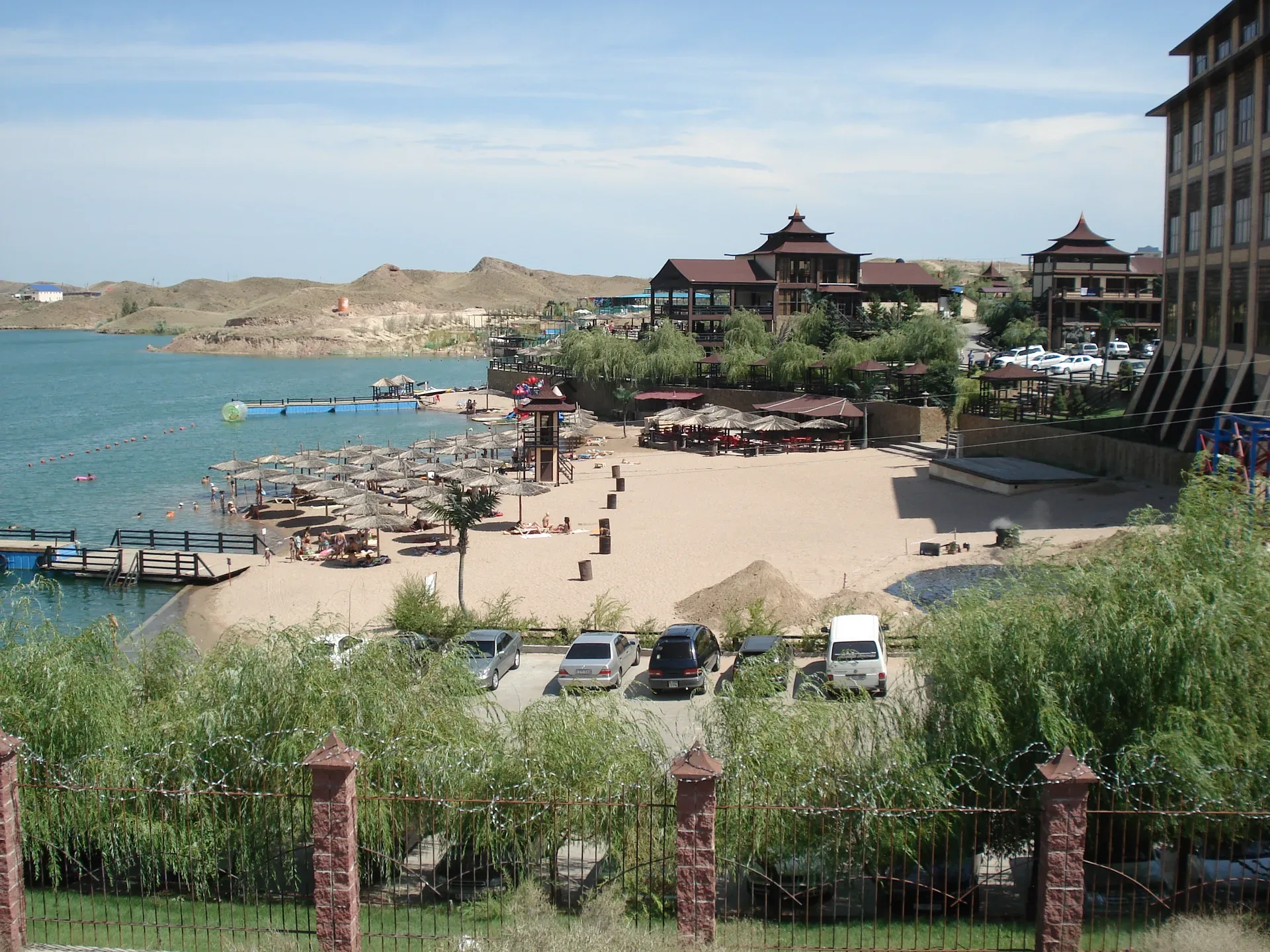 Kapchagai reservoir shoreline in Kazakhstan, calm blue-grey water with low sandy banks and sparse vegetation under hazy sky