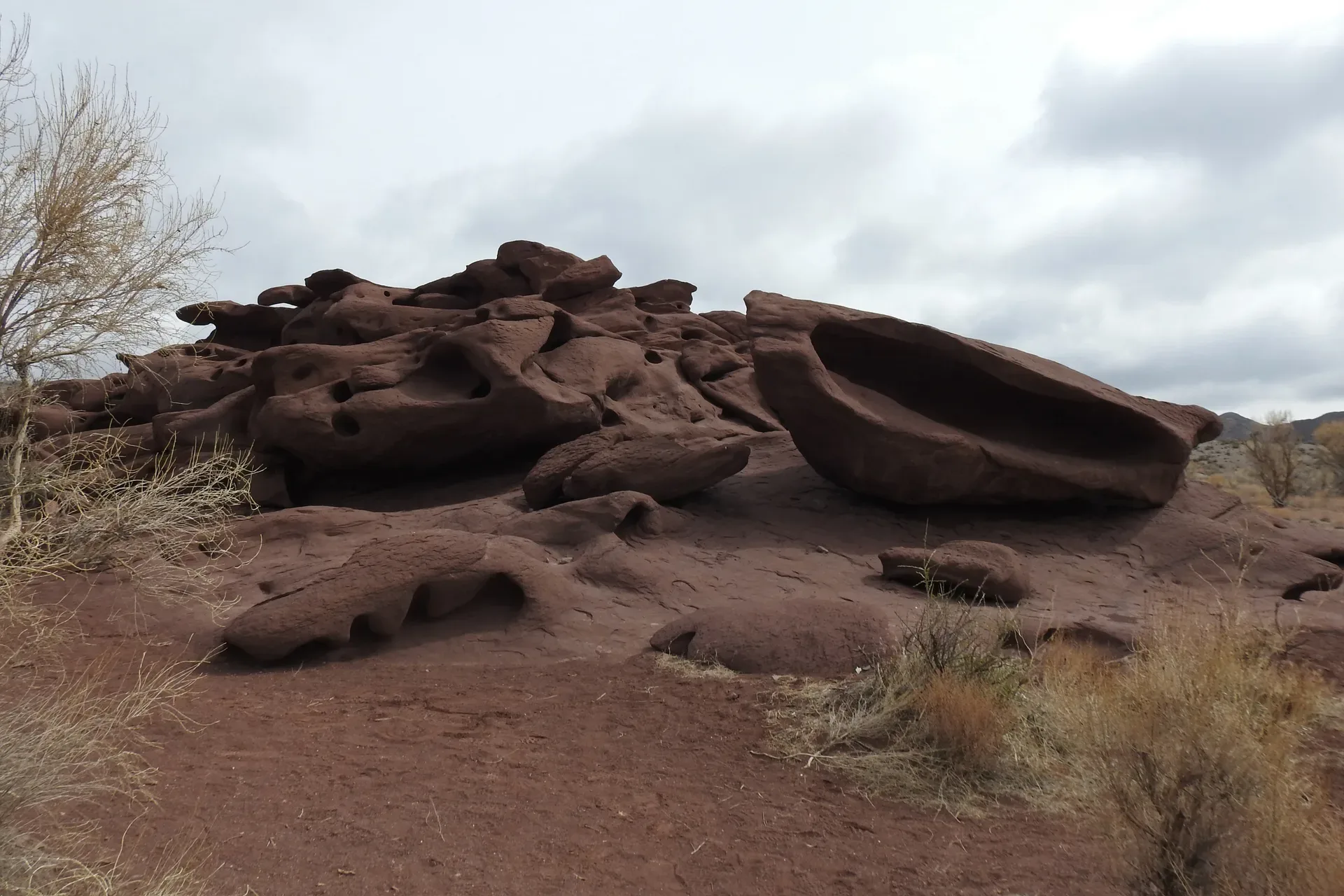 Eroded dark reddish-brown volcanic rock formations of the Katutau Mountains in Altyn-Emel National Park, showing windswept solidified lava outcrops rising above desert scrub under an overcast sky.