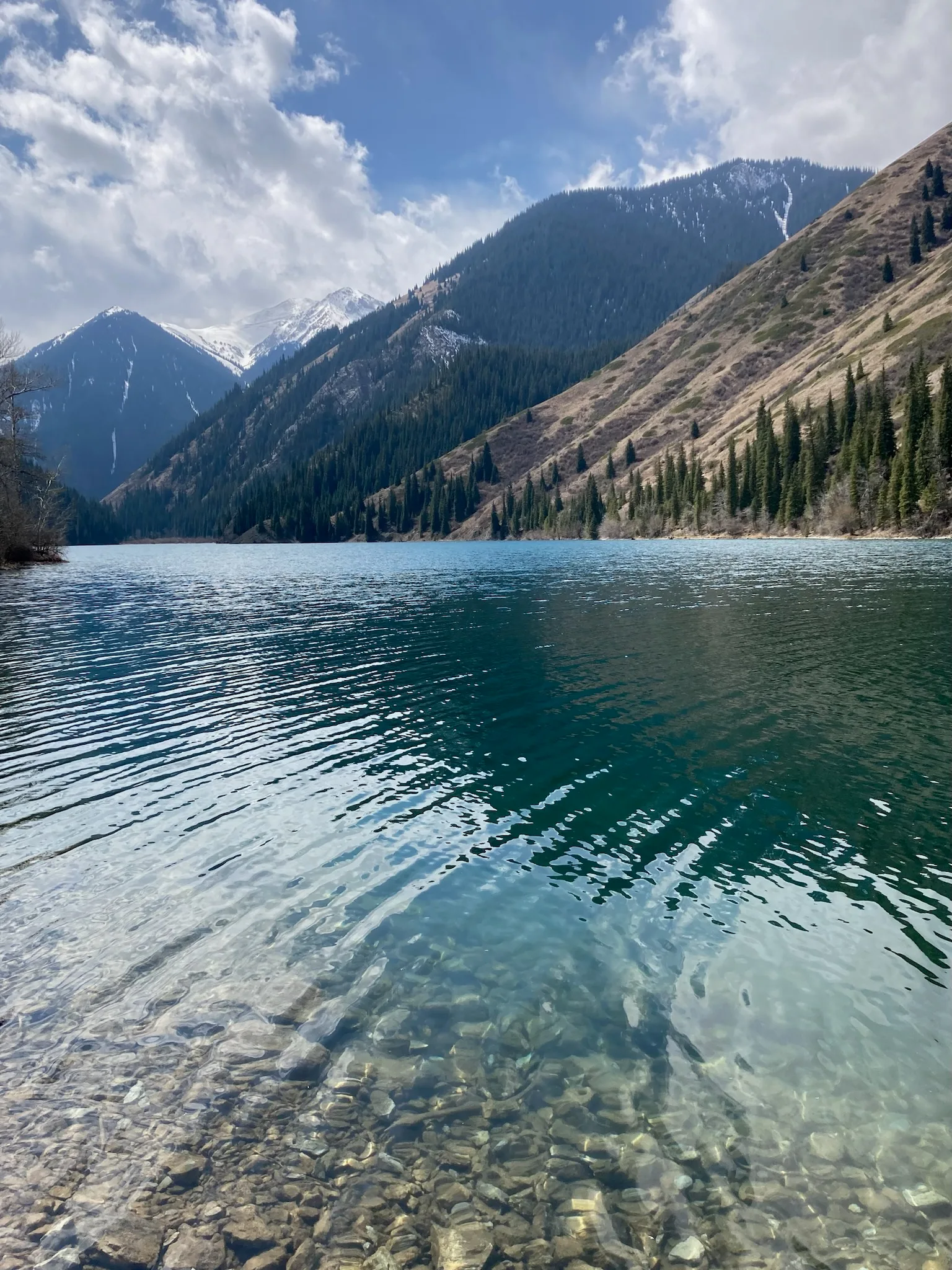 Lower Kolsai Lake in April, with cold blue water, bare shoreline vegetation, and forested mountain slopes.