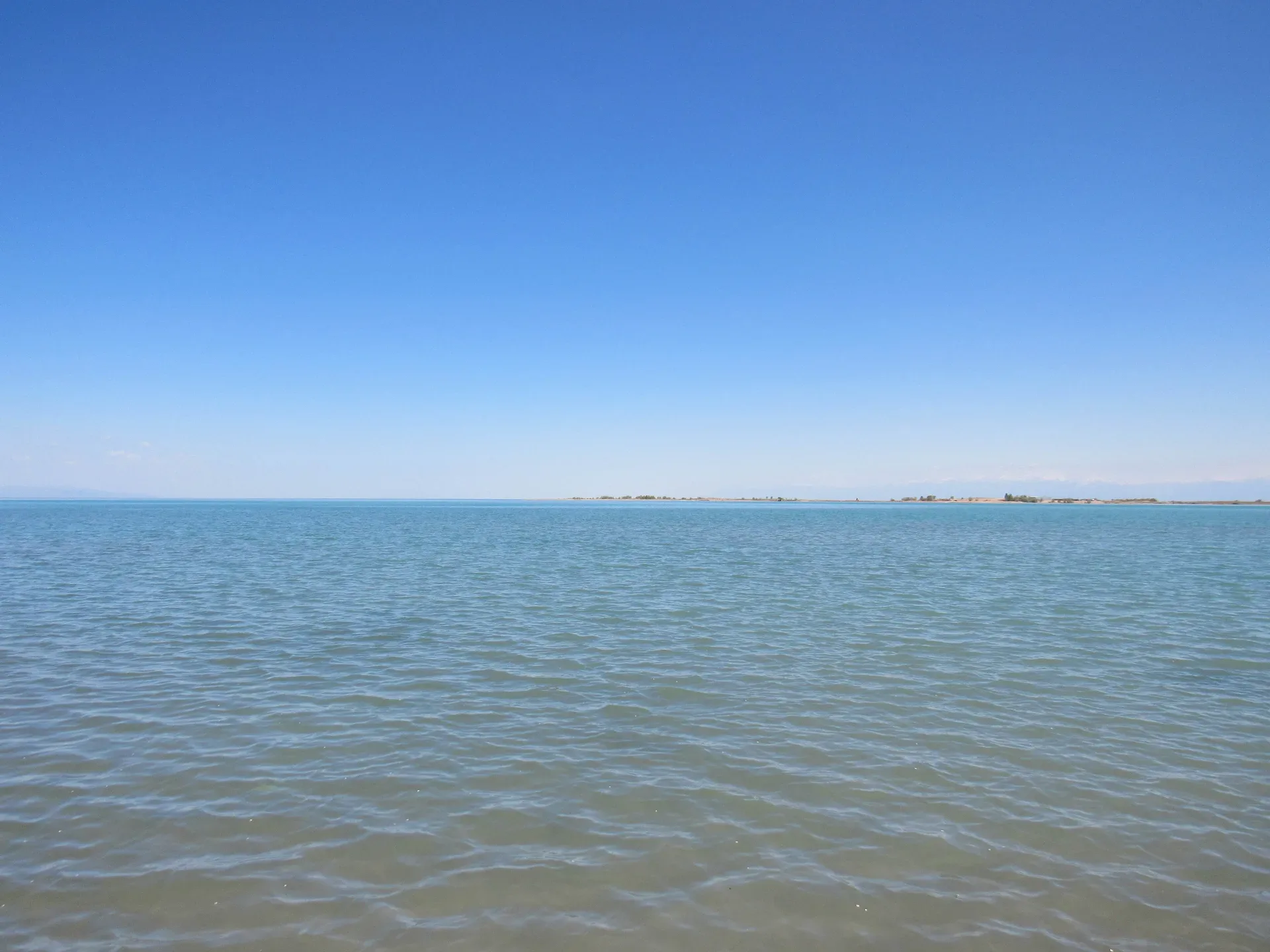 Kapchagay reservoir shoreline at Konaev city, calm blue water with a gravel bank in the foreground under a pale grey sky