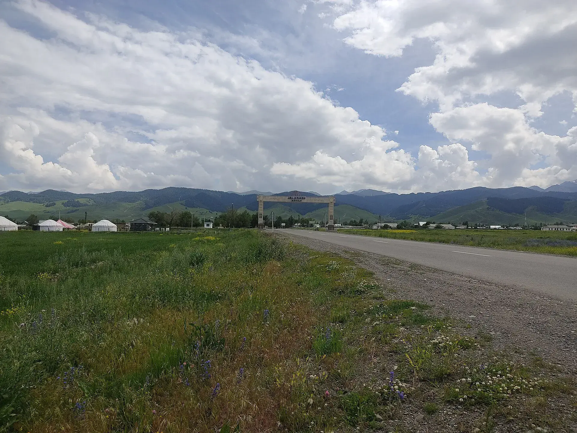 Entry road to Zhalanash village with flat Kegen steppe and low hills beyond the road edge.