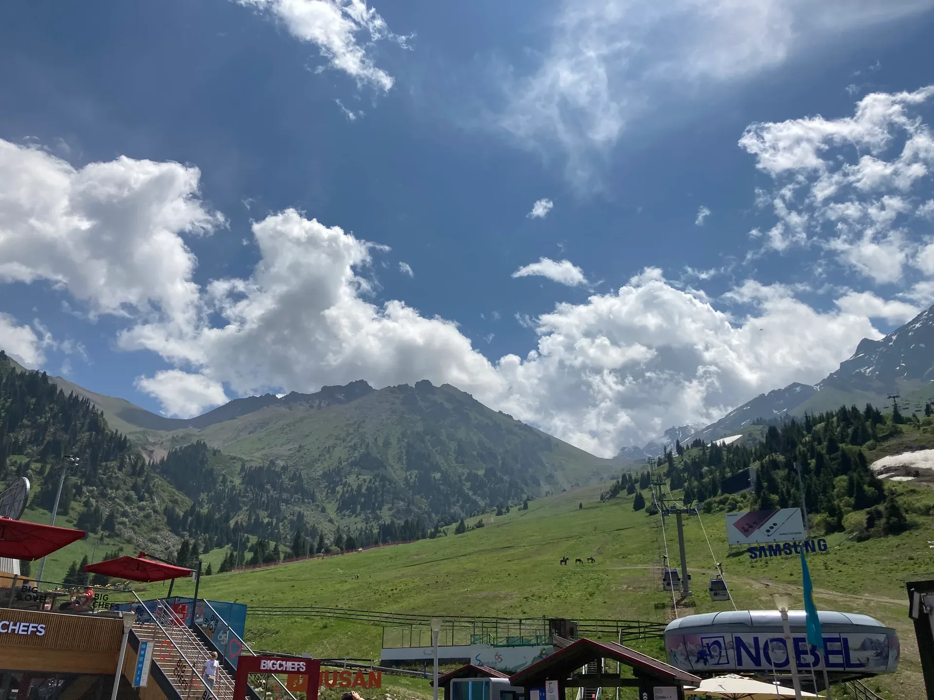 Shymbulak ski resort base station at 2,260 m in summer, with gondola terminal buildings, chairlift infrastructure, restaurant terraces, and green alpine meadows backed by the Zailiisky Alatau peaks.