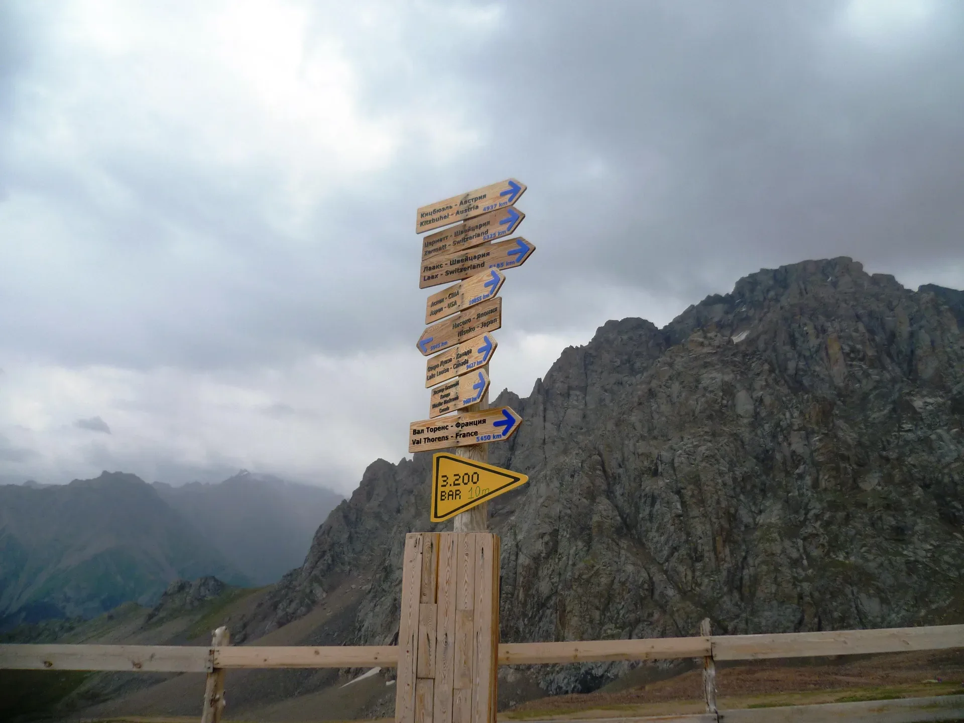Direction signs at Talgar Pass in the Shymbulak area, painted wooden column with trail distances, rocky alpine terrain in background