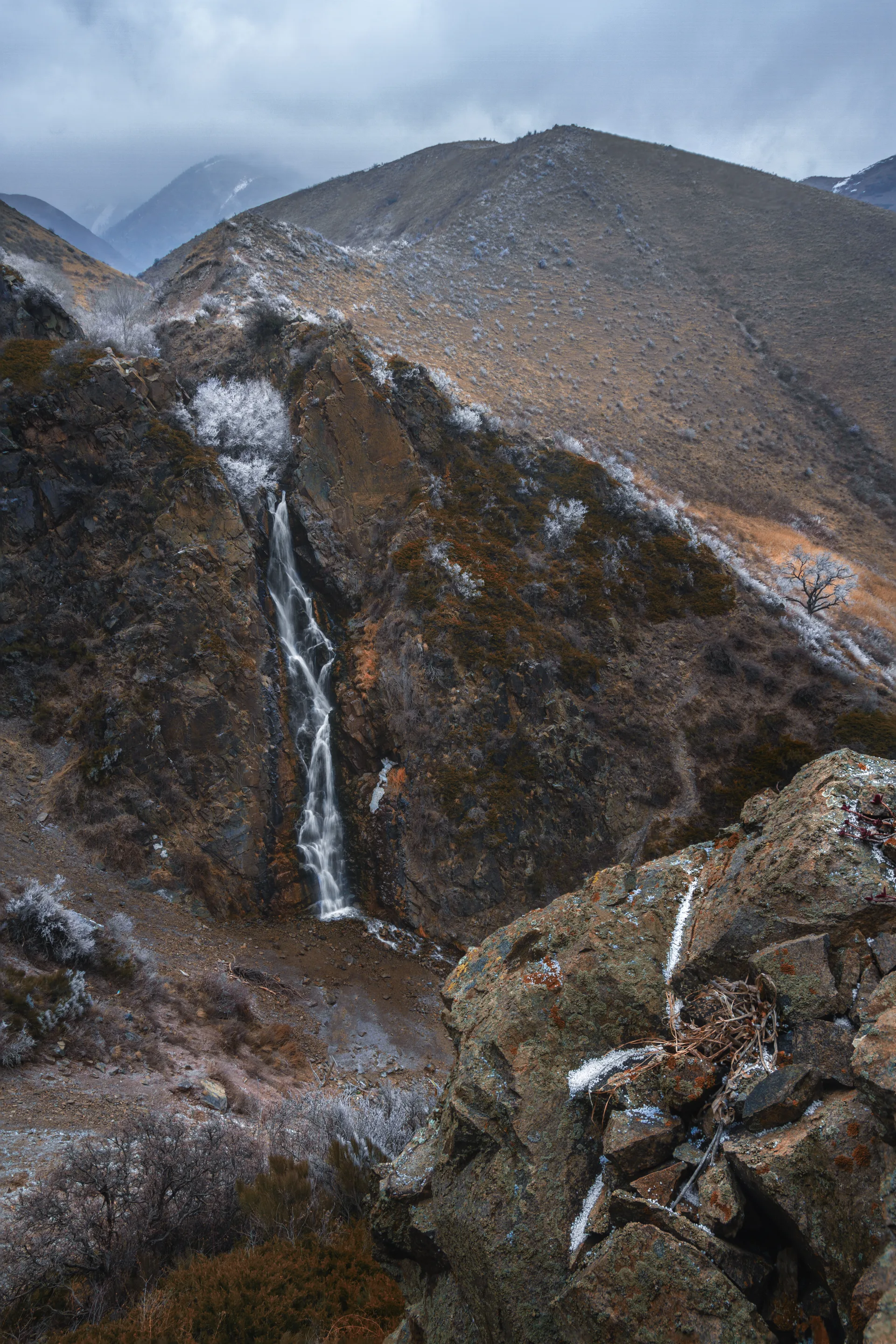 Medvezhiy (Bear) Waterfall in Turgen Gorge falling 30 metres into a rocky basin surrounded by moss-covered boulders and conifer forest.