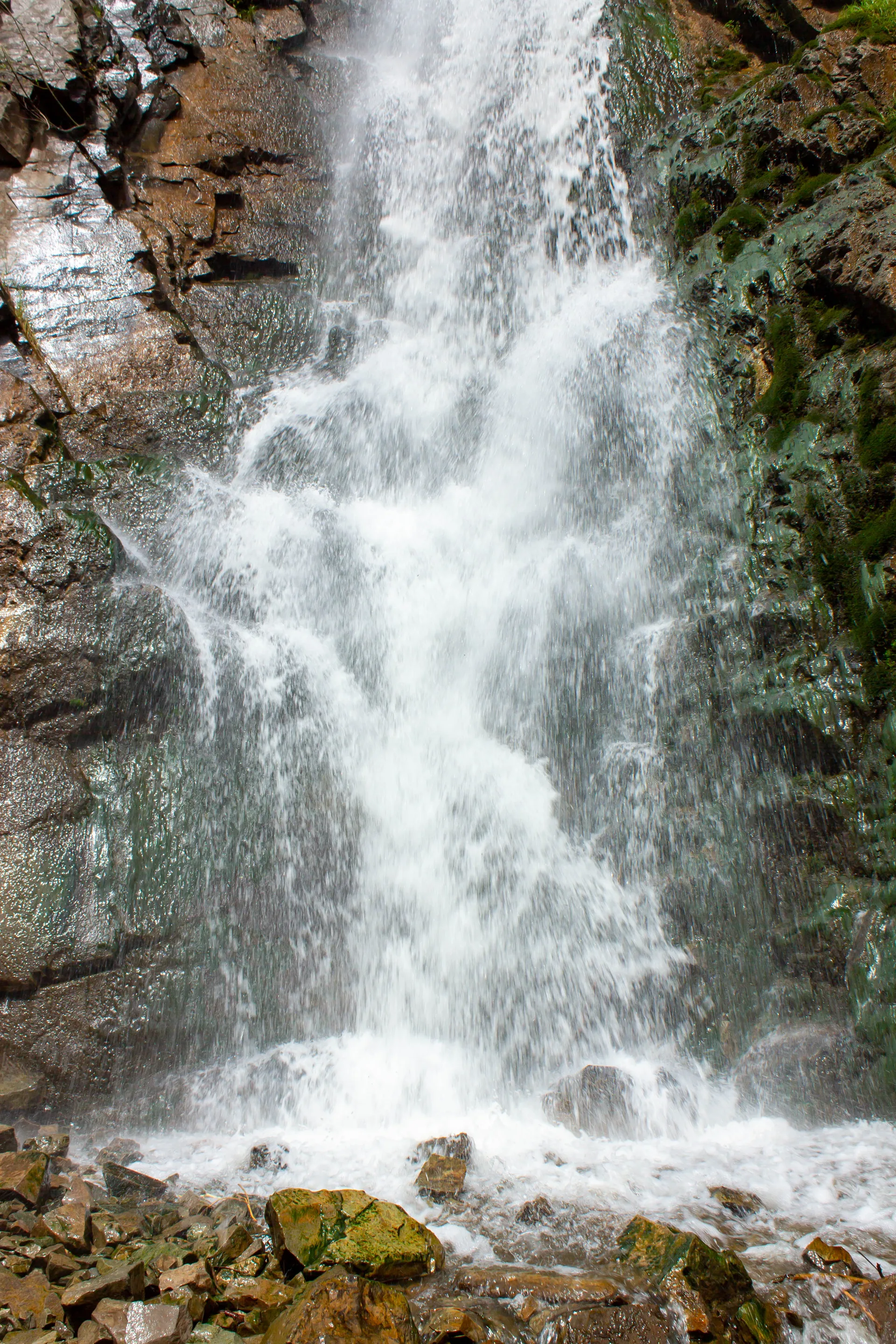 Medvezhiy (Bear) Waterfall in Ile-Alatau National Park cascading over rock with mist and moss-covered boulders at the base.