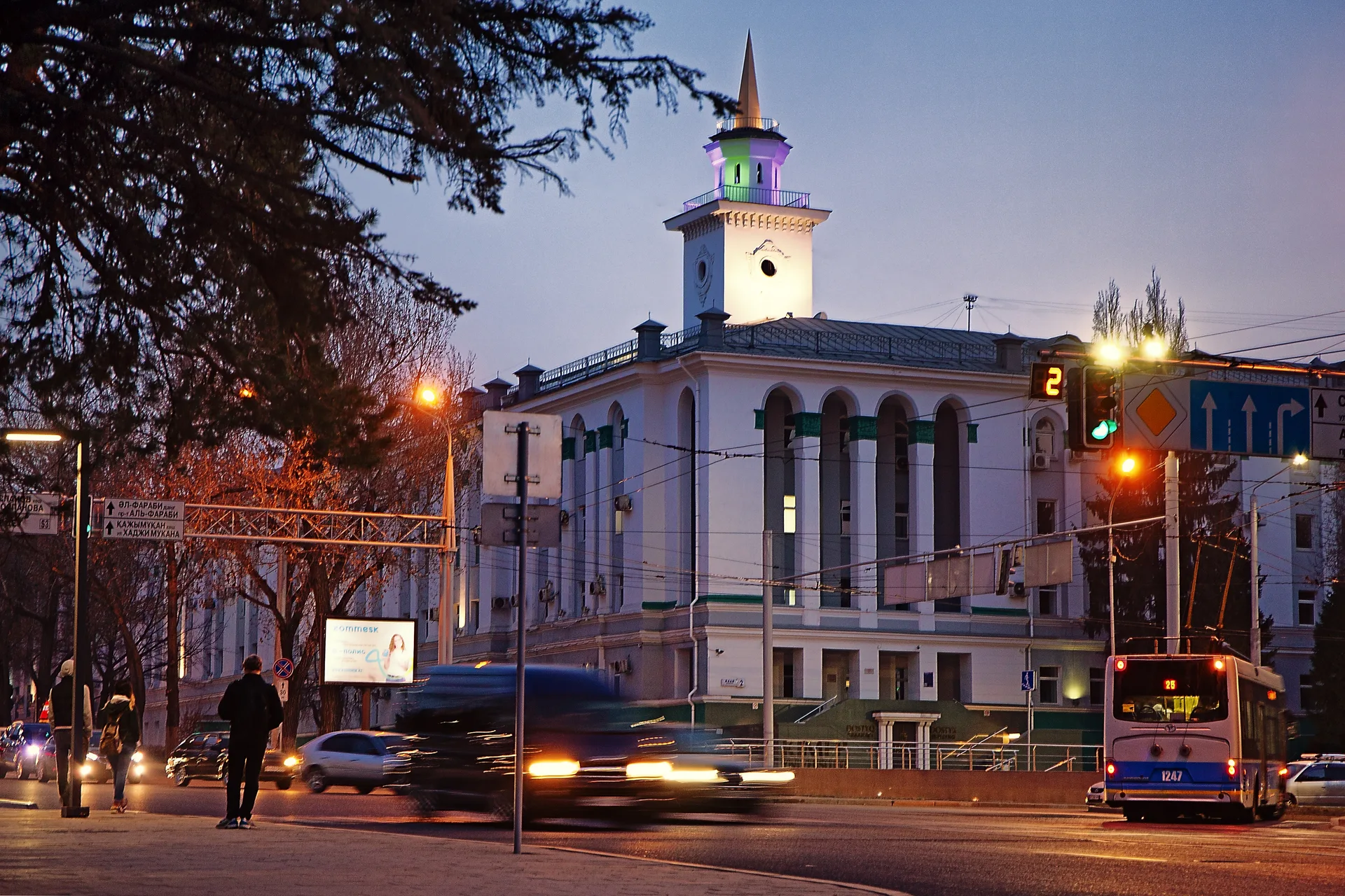 Dostyk Avenue in Almaty at dusk, wide tree-lined boulevard heading south toward the mountains with headlights and traffic