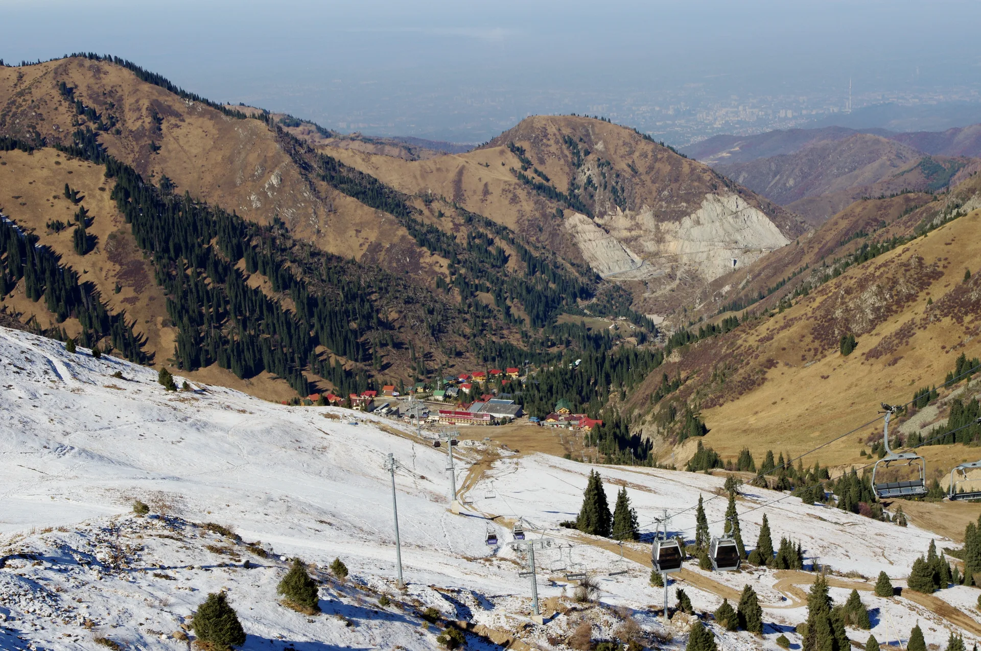 View north from Shymbulak ski slopes toward Almaty city, autumn colours on lower slopes with urban grid and steppe visible in the far distance