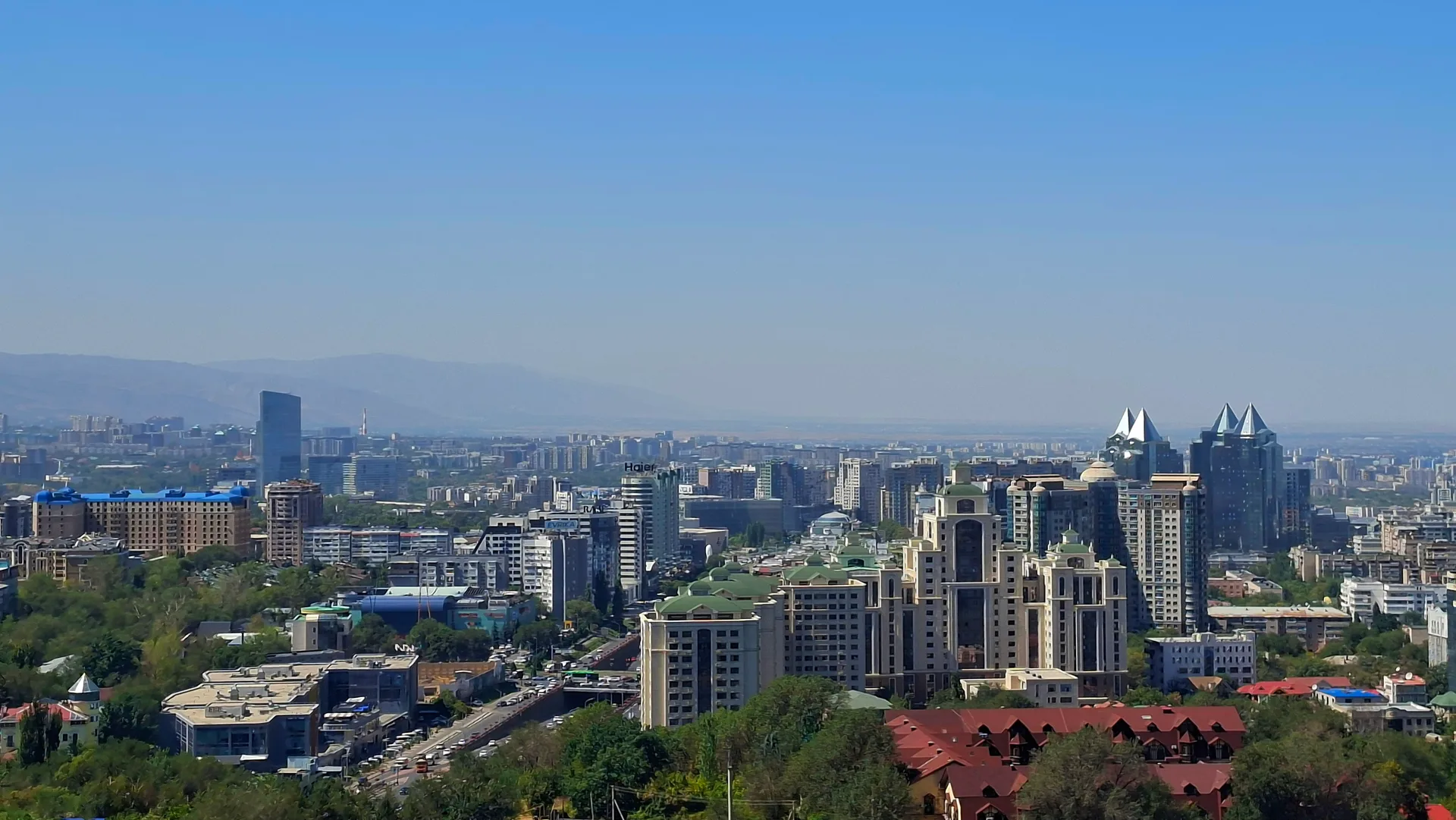 Almaty city skyline seen from Kok Tobe hill, mid-rise buildings and tree-lined streets in the foreground, Zailiysky Alatau mountain range behind